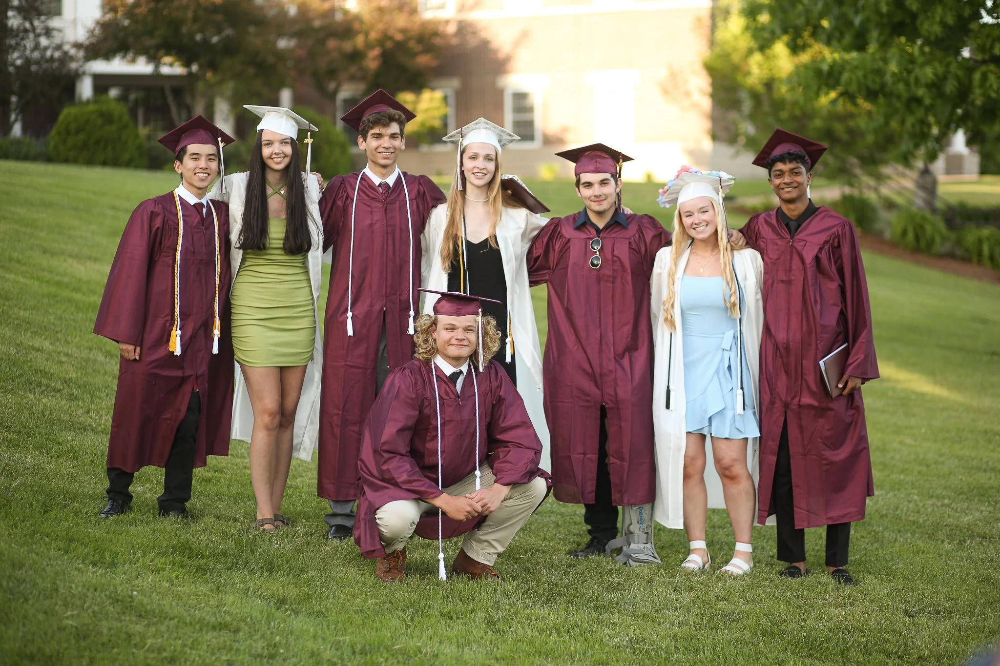Group of diverse young adults dressed in graduation caps and gowns, standing on grass outdoors during daytime.