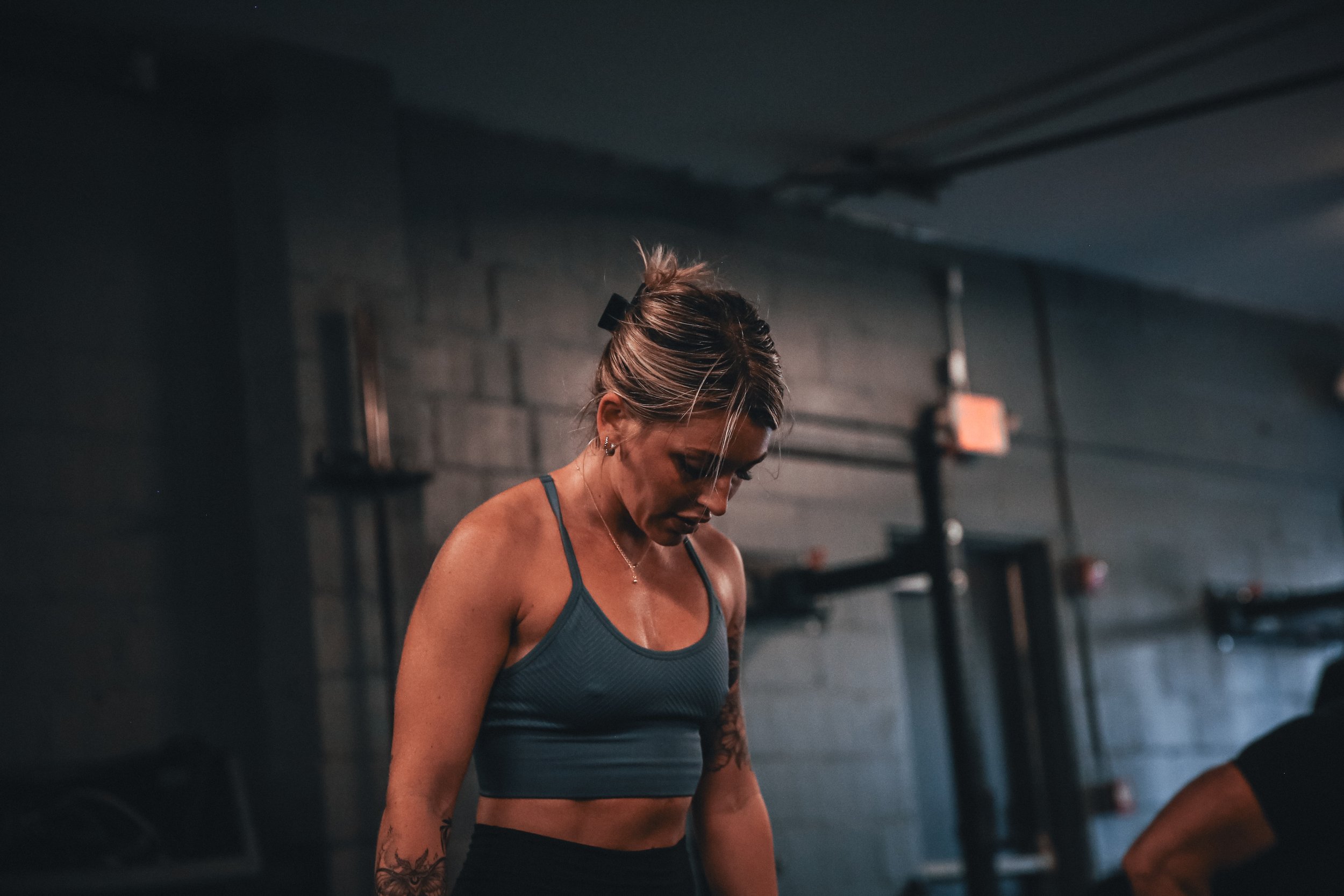 A woman in workout clothes in a gym, looking down, with a dark background and gym equipment.