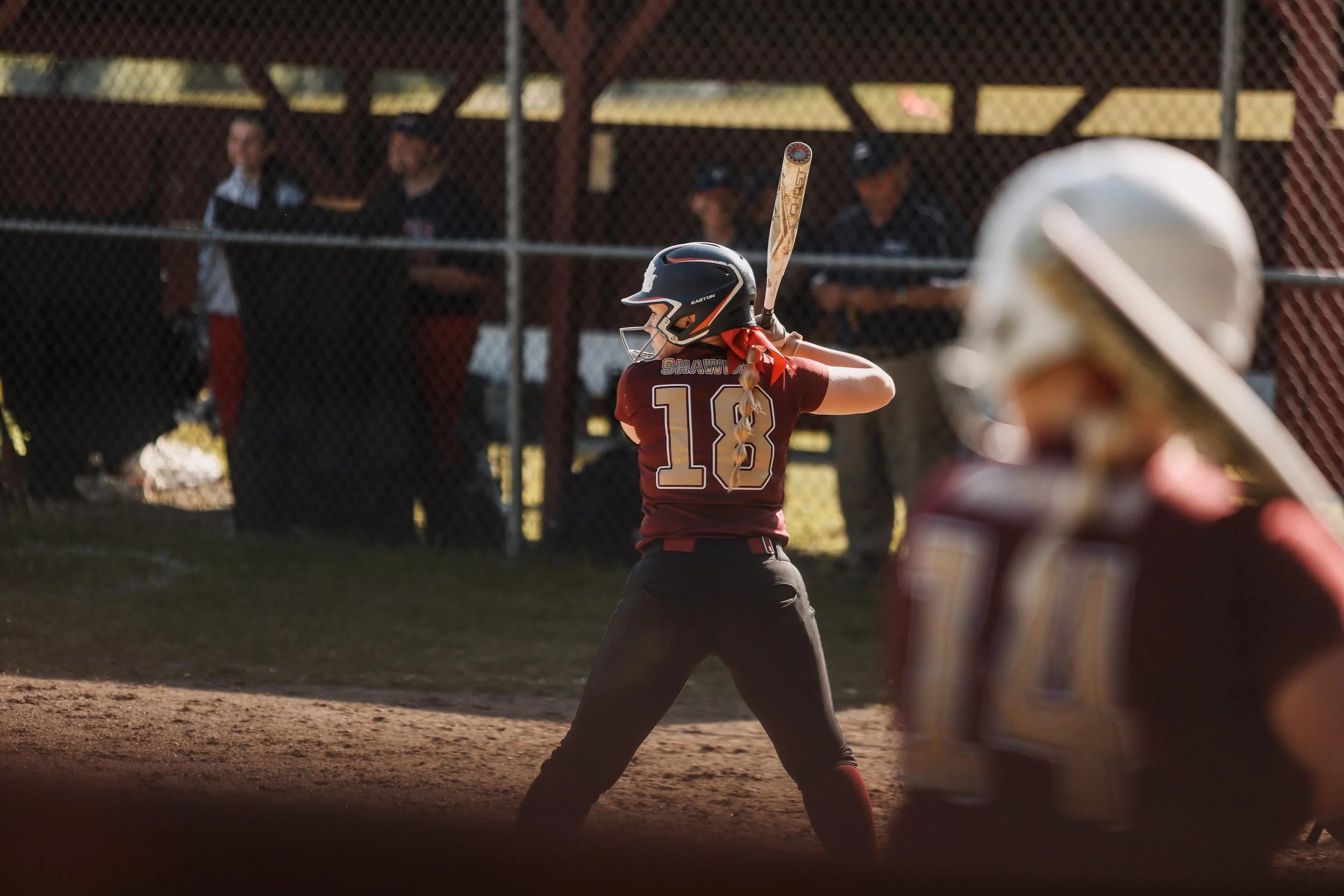 Youth baseball player in maroon uniform and helmet preparing to swing bat on the field, with a catcher in the foreground and spectators behind a chain-link fence in the background.