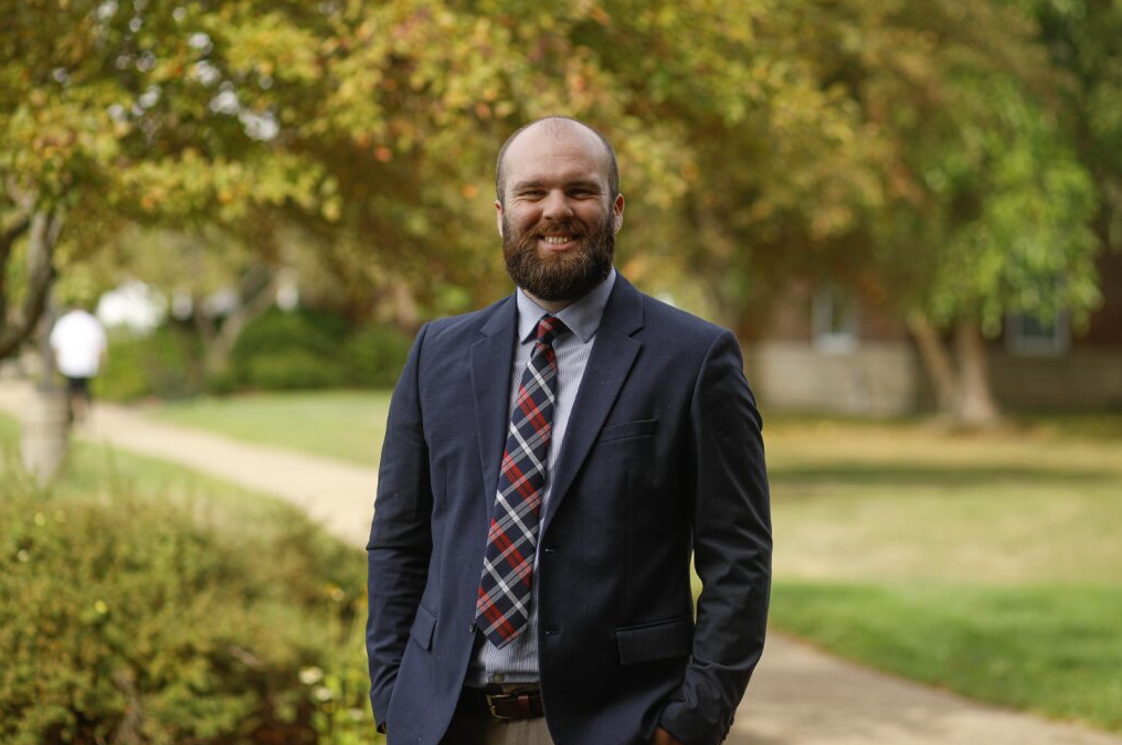 A man in a jacket and patterned tie standing outdoors on a sidewalk, smiling, with trees and a building in the background.