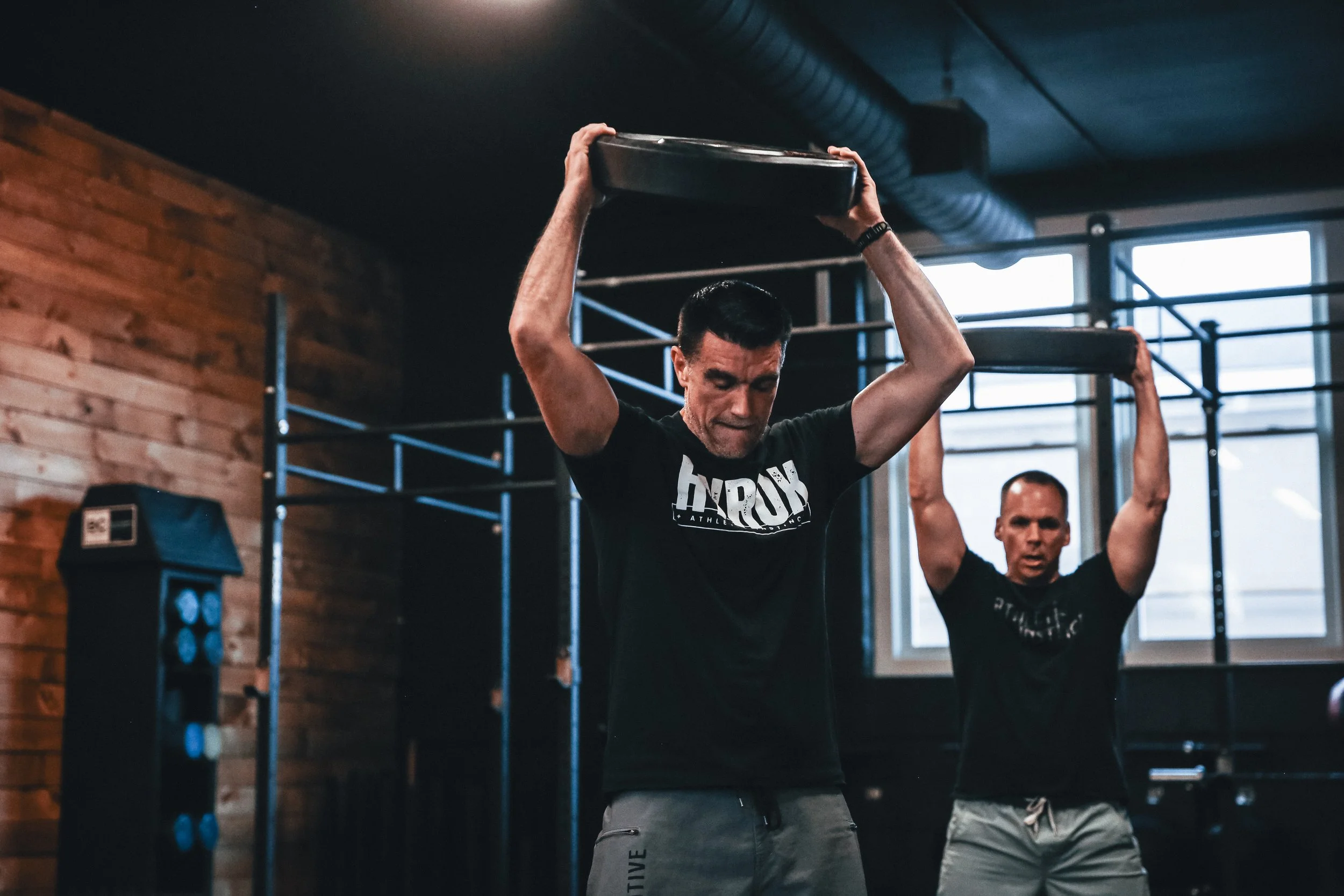 Two men lifting weight plates during crossfit training in a gym with wooden walls and large windows.