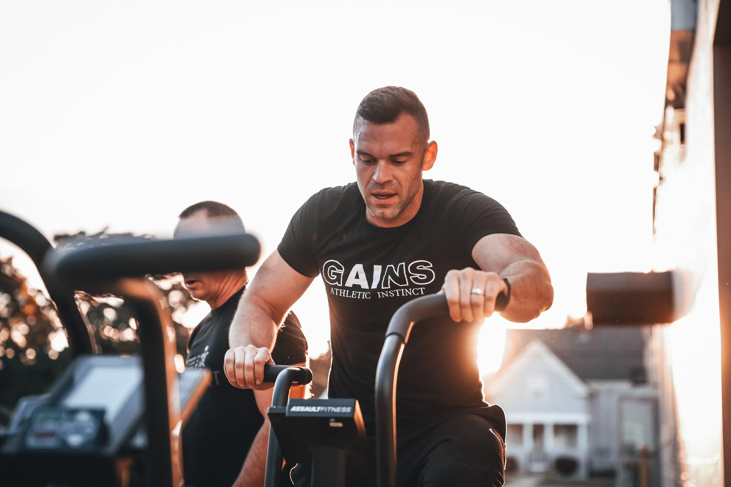 Two men are exercising outdoors at sunset, riding stationary bikes. The man in the foreground is wearing a black t-shirt with the words 'GAINS ATHLETIC INSTINCT' and has a focused expression.