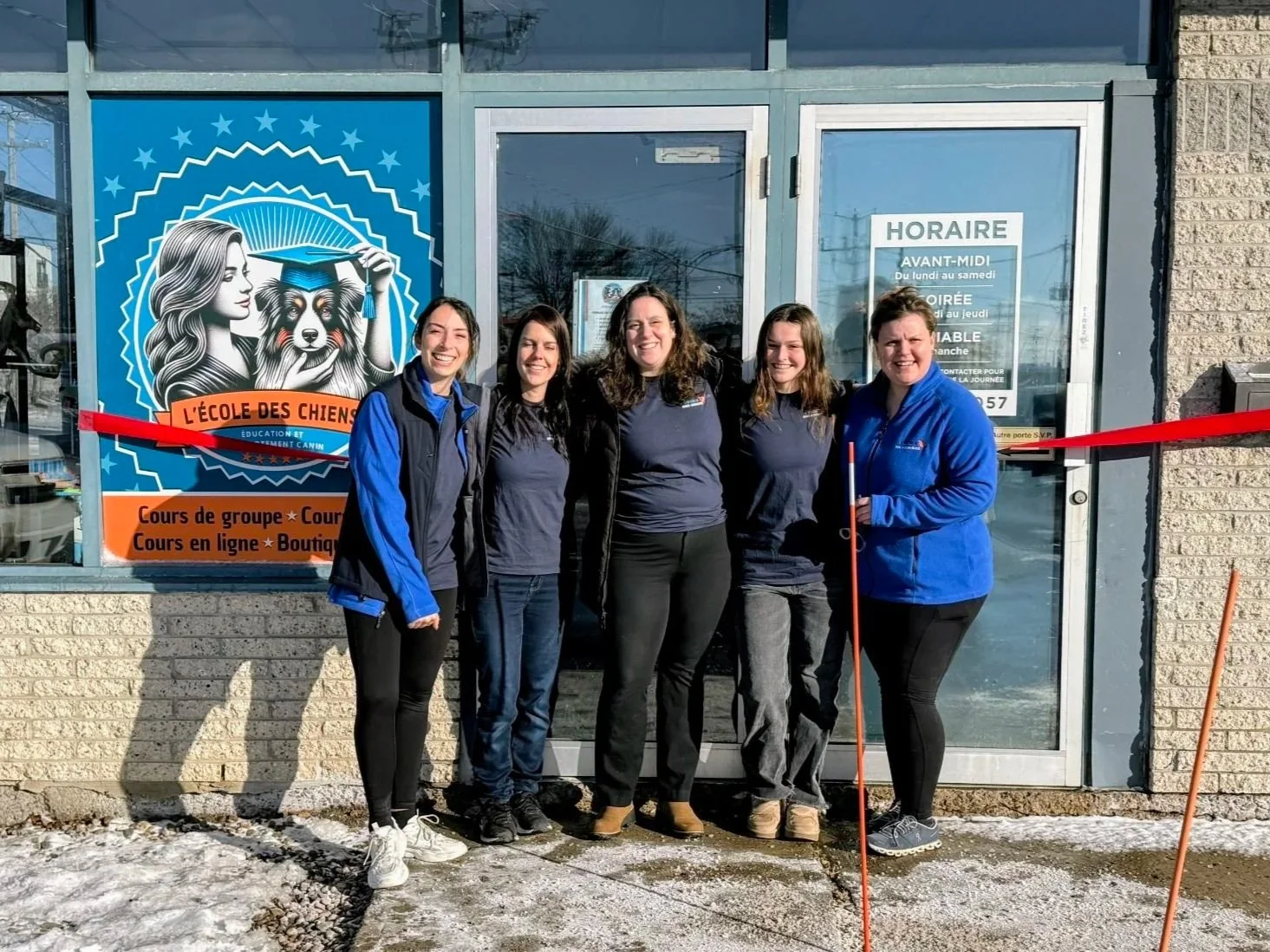 Cinq femmes debout devant la vitrine d'une école pour chiens, souriantes, avec une bande rouge de ruban de coupe en bas. La vitre affiche un logo avec une femme, un chien et le texte "L'école des chiens".