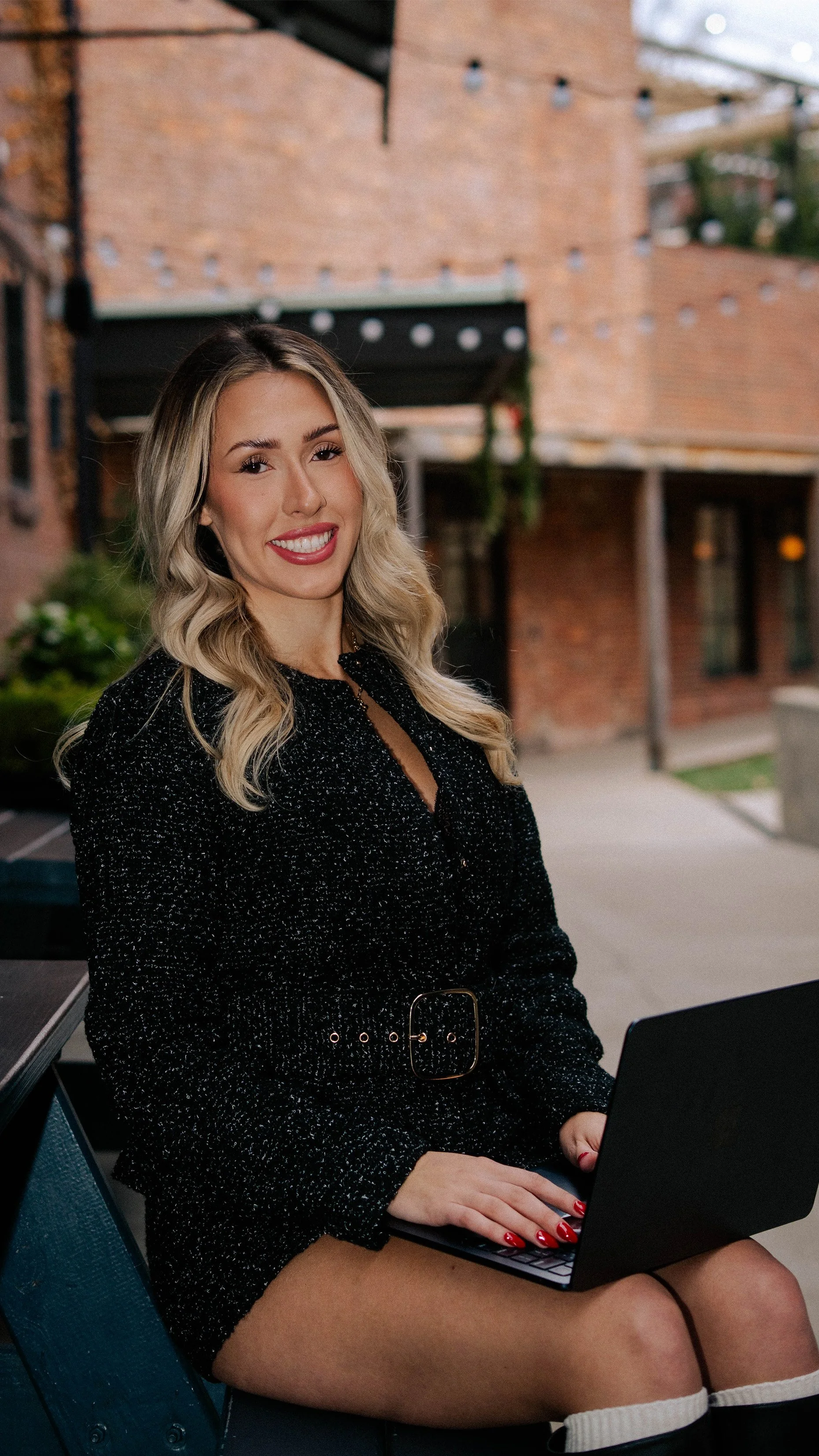 A woman sitting outdoors at a table with a laptop, smiling with a brick building and string lights in the background.