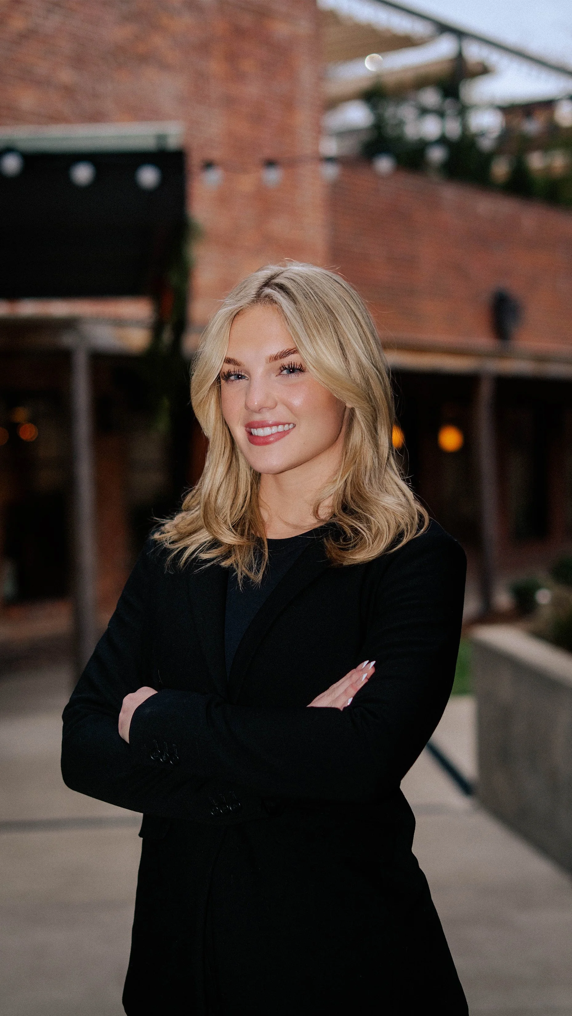 A woman with blonde hair wearing a black blazer, smiling with her arms crossed outside in front of a brick building.