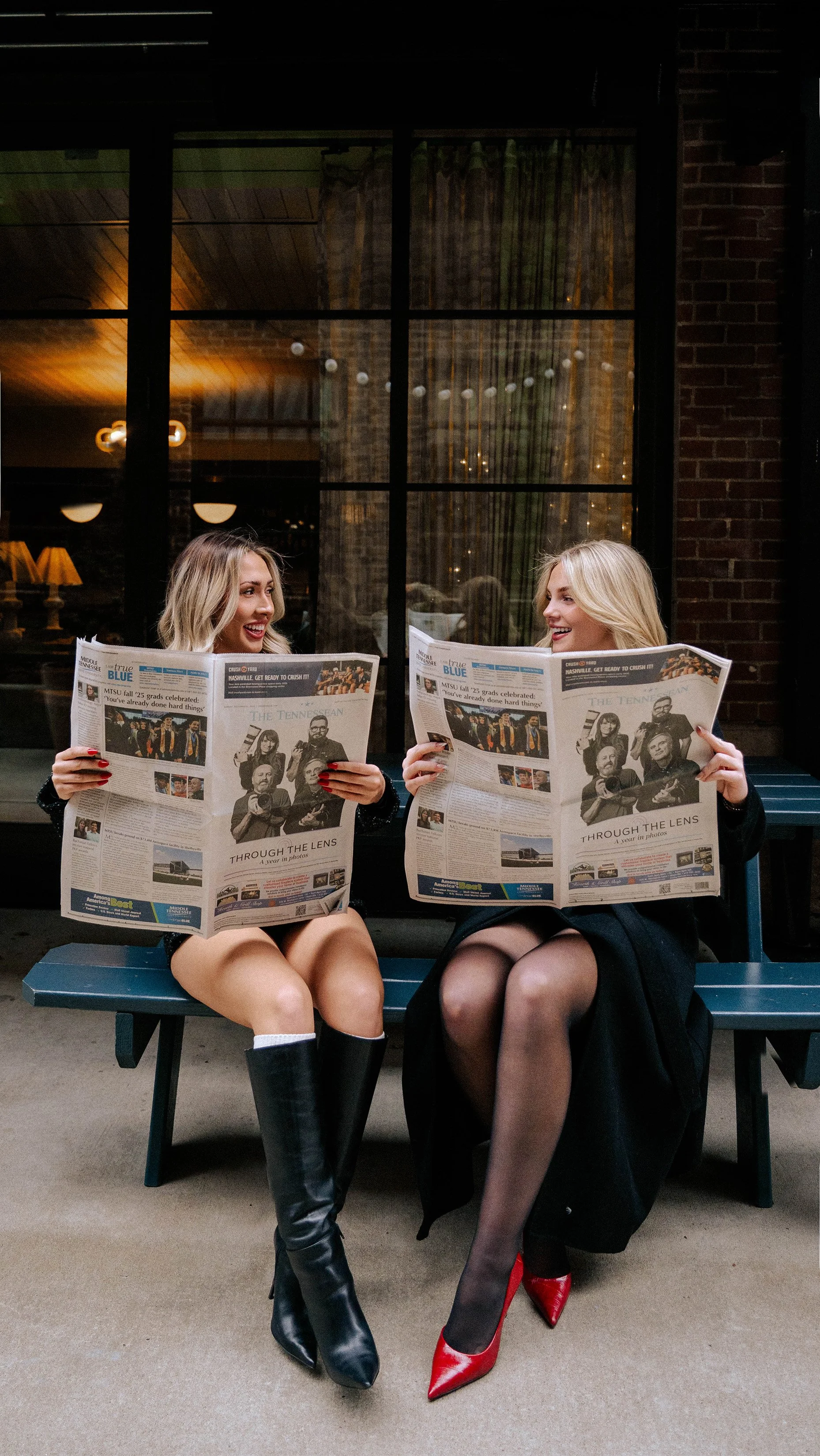 Two women sitting on a bench outside under a shelter, smiling and talking while reading newspapers, with a large window and warm indoor lighting in the background.