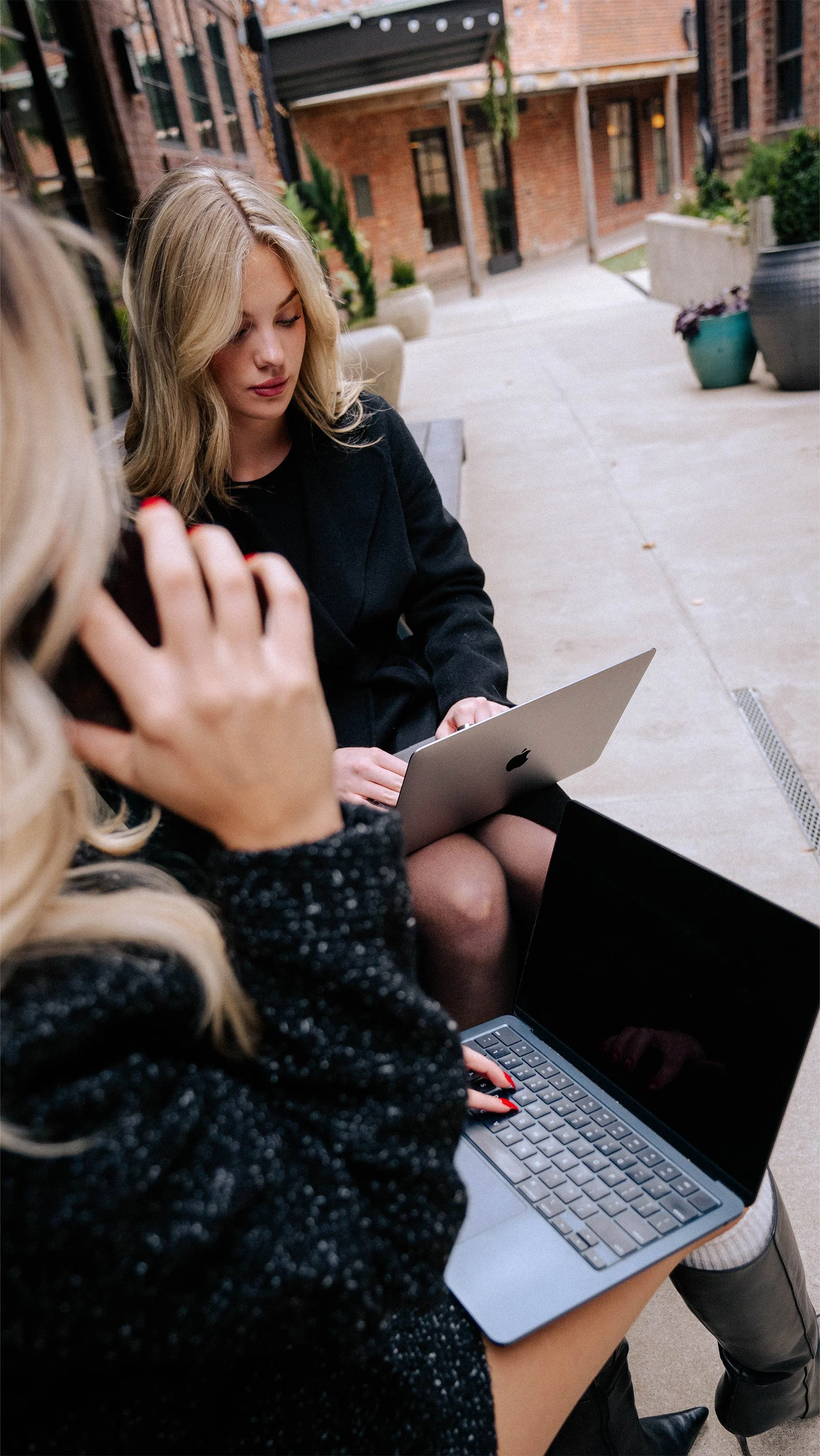 Two women with blonde hair working on laptops outdoors on a patio with potted plants and brick buildings in the background.