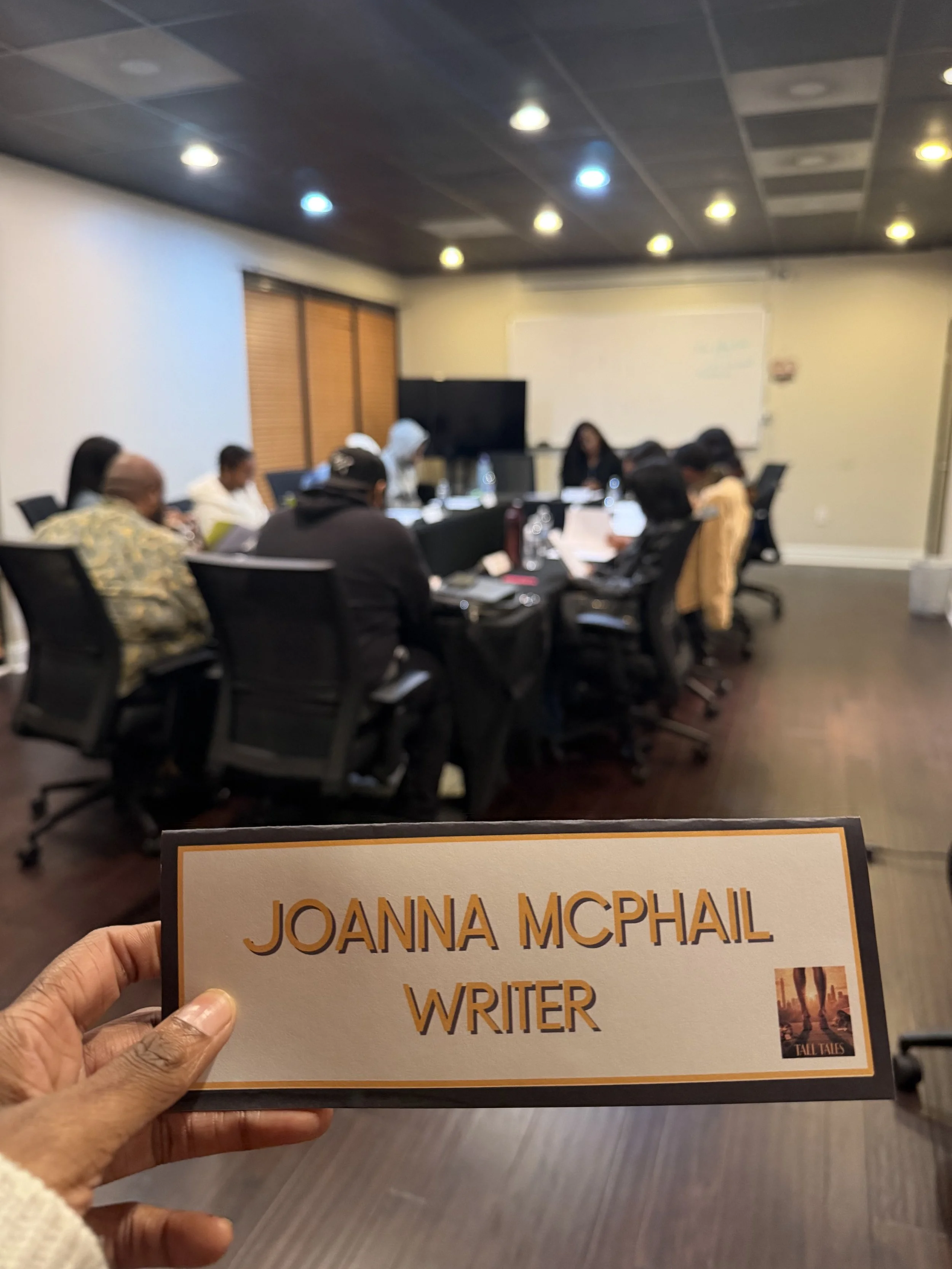 A person holding a nameplate with the text 'JOANNA MCPHAIL WRITER' in front of a blurred conference room with several people seated around a table.