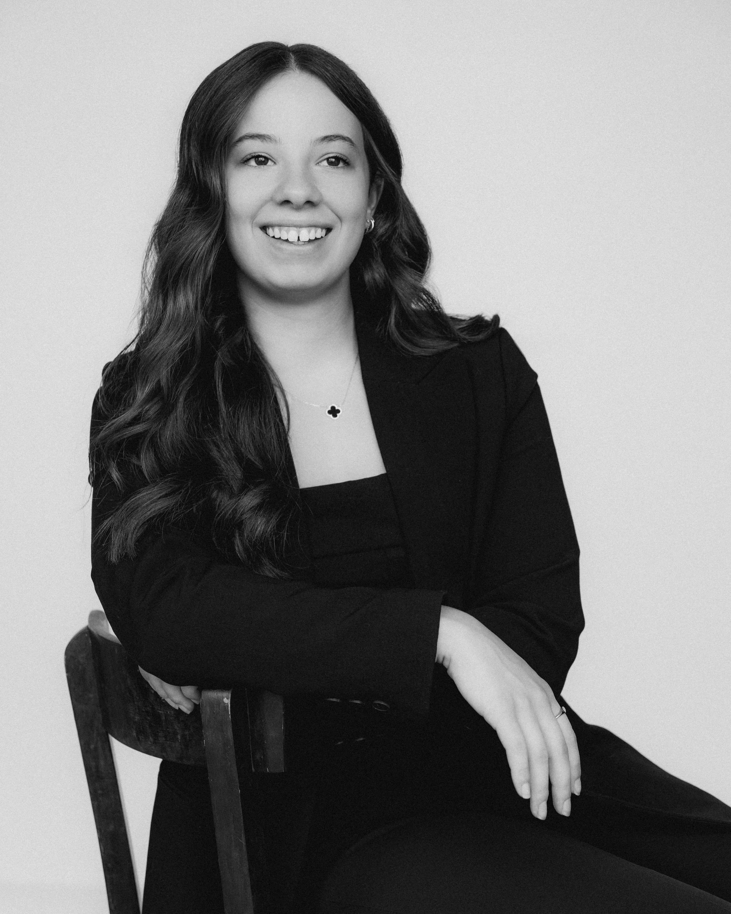 Portrait en noir et blanc d'une femme souriante assise sur une chaise, portant un blazer noir, une chemise noire, et un collier avec un pendentif en forme de trèfle.