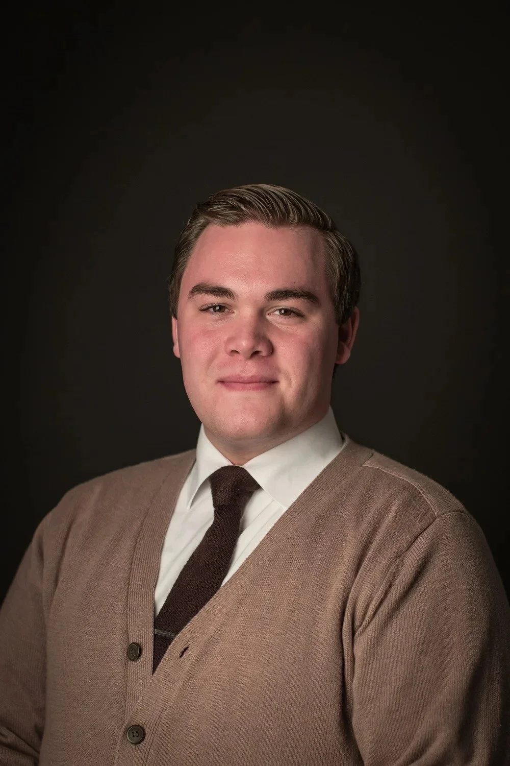 A young man with styled hair in a brown cardigan, white shirt, and dark tie, posing against a dark background.