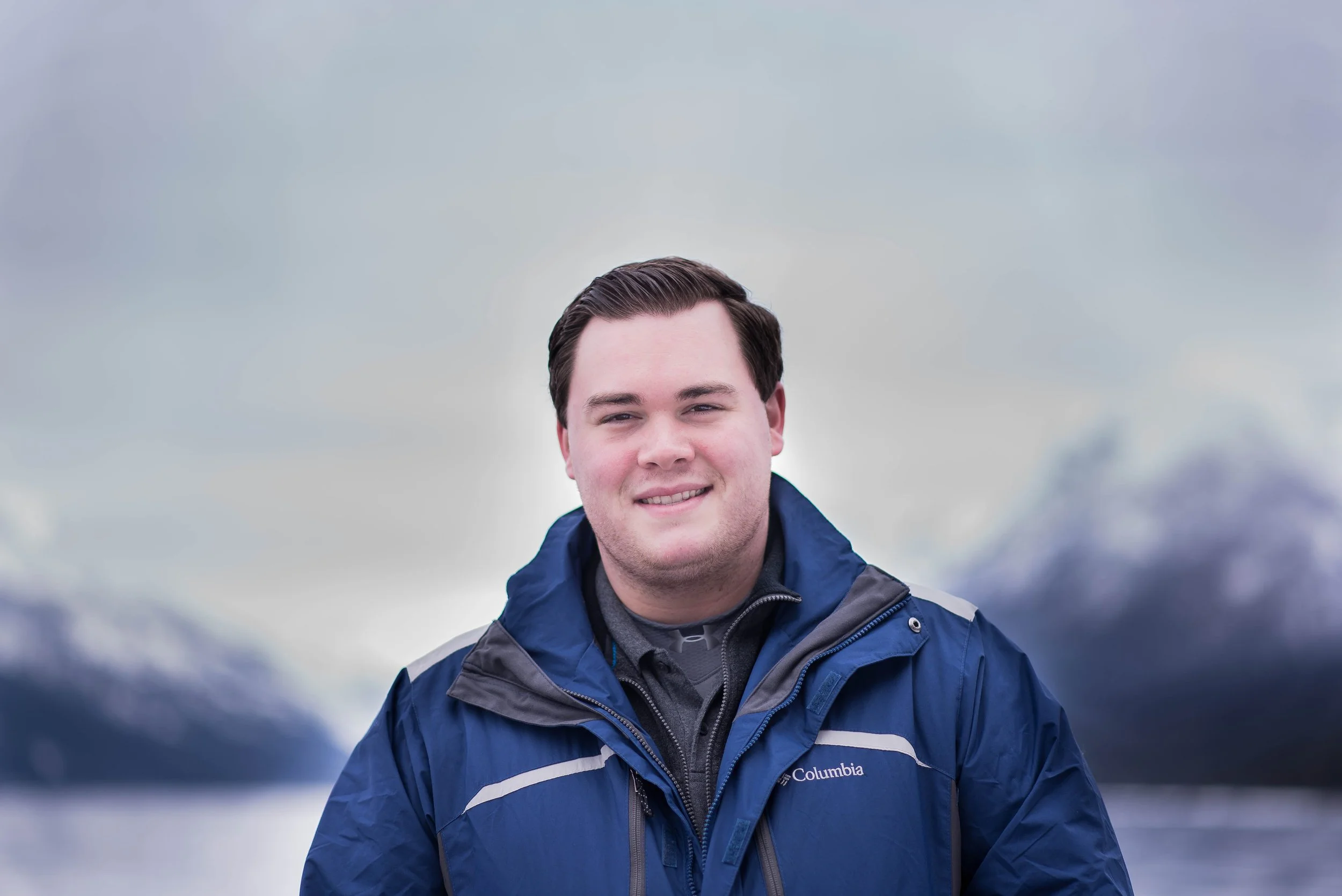 A young man with short dark hair smiling at camera, wearing a blue Columbia jacket, standing outdoors with mountains and water in the background.