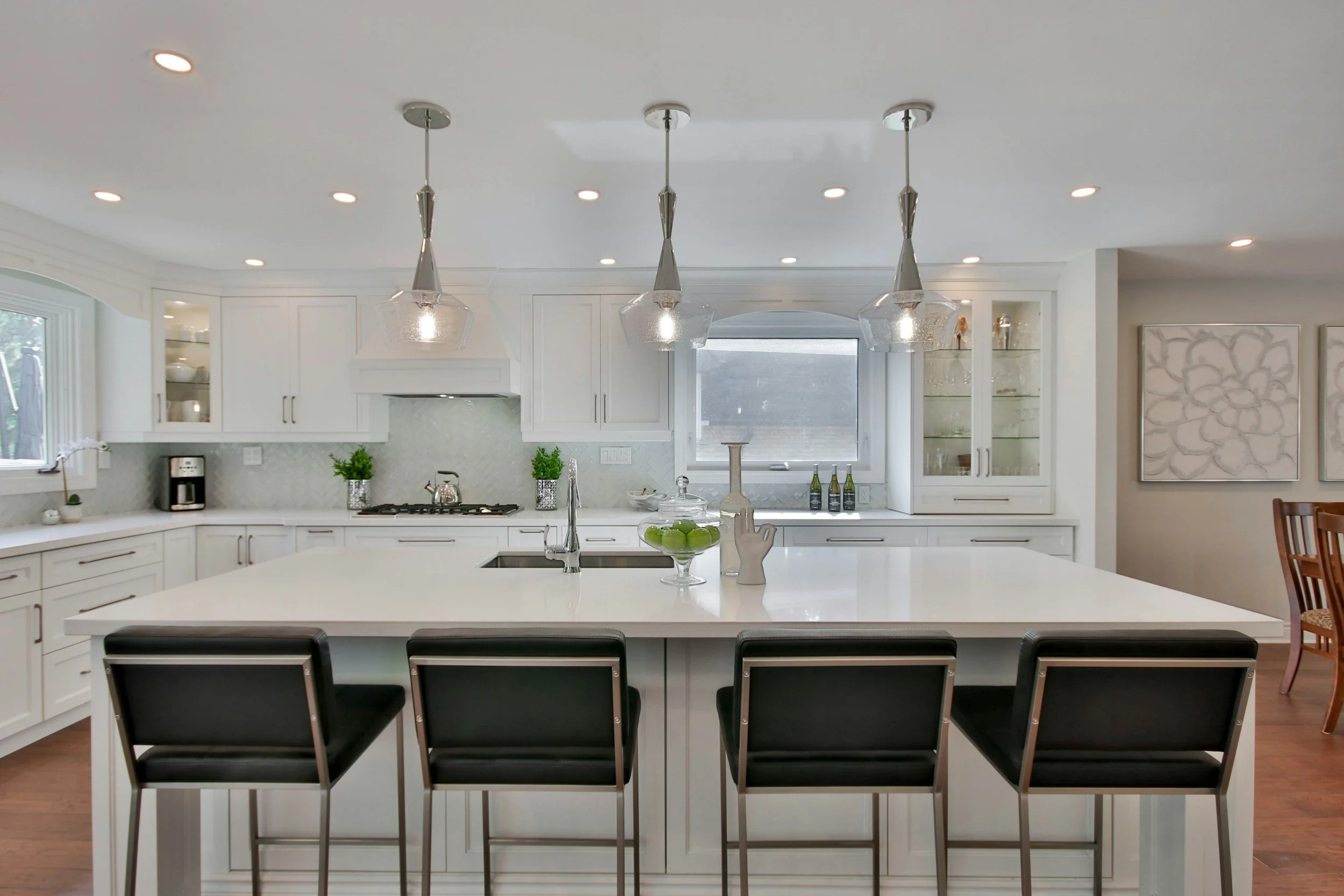 Modern white kitchen with large island, black bar stools, pendant lights, and white cabinetry.