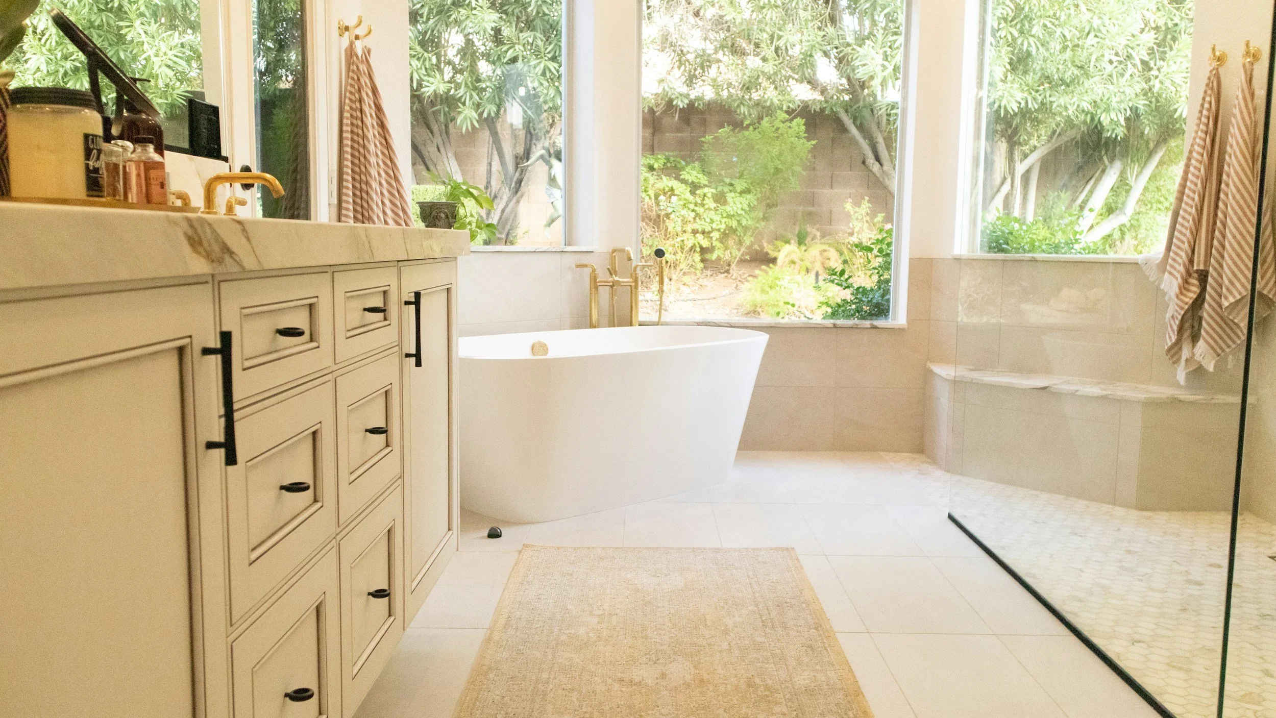 Bright bathroom with large windows overlooking greenery, featuring a white bathtub, beige tiled floor, a cream-colored cabinet with black handles, and a glass-enclosed shower area with pebble tiles.