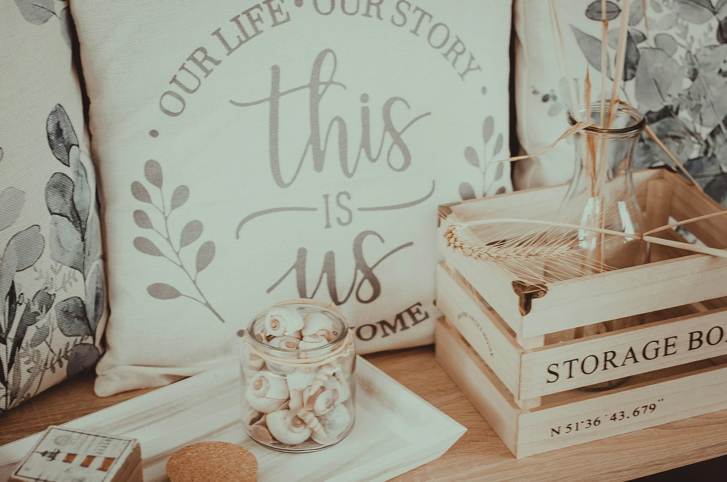 Decorative pillows, a glass jar filled with seashells, a small cork coaster, and a wooden box labeled 'Storage Box' with a glass vase and a feather inside sit on a wooden surface. The background features a pillow with the text 'This is us' and floral patterns.