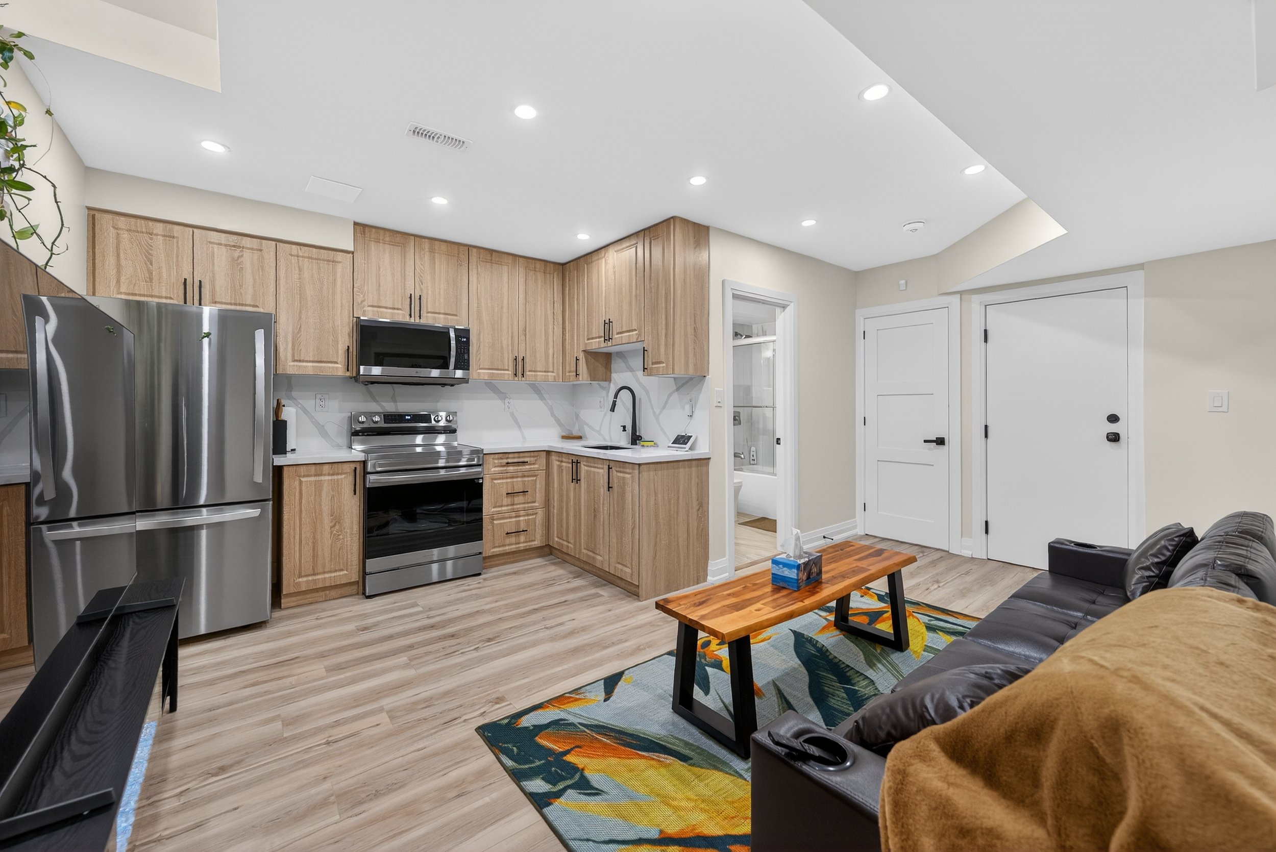 Modern open-concept living room and kitchen with wood cabinets, stainless steel appliances, black faucet, wooden coffee table, black leather couch, colorful patterned rug, and hardwood floors.