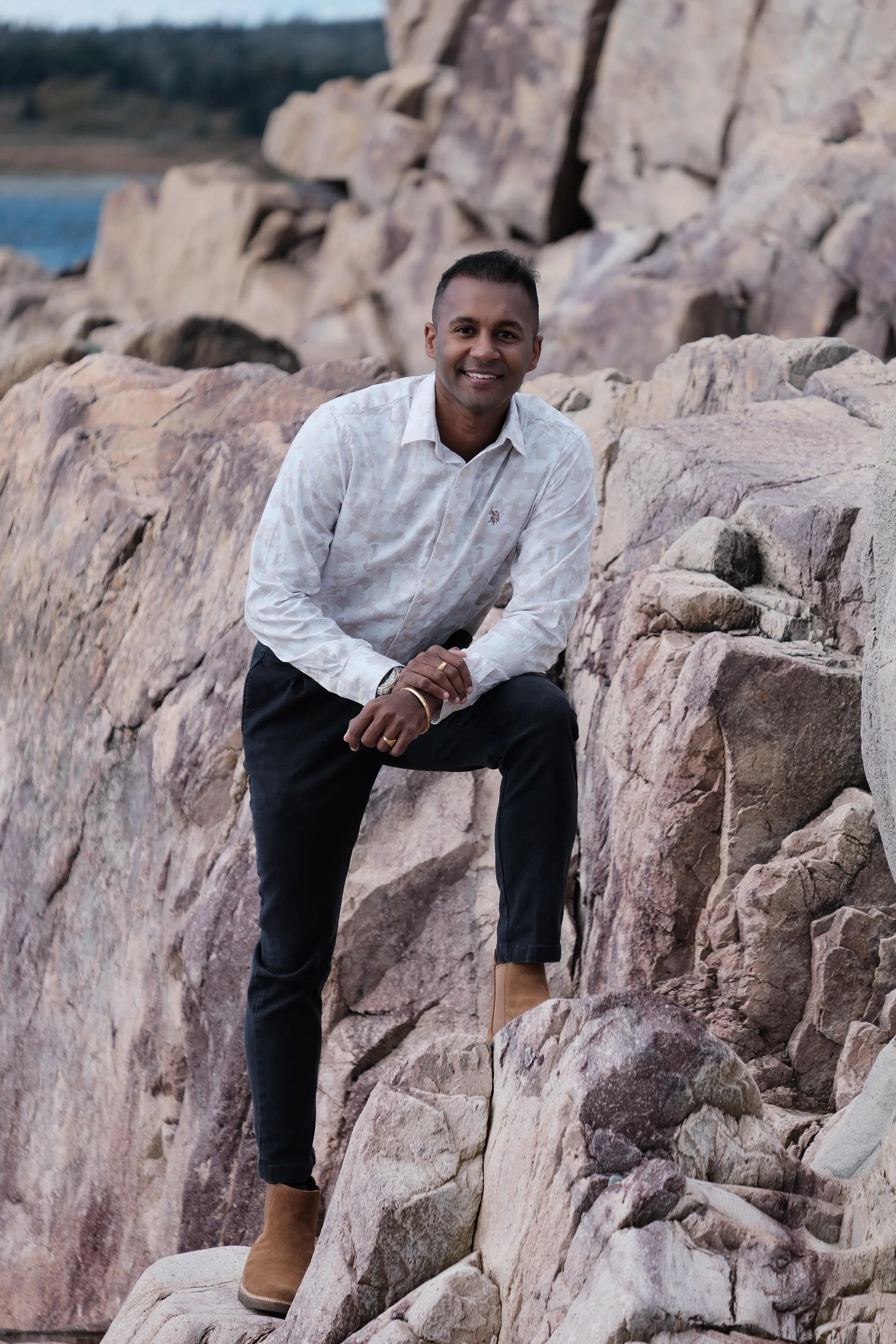 A man in a white patterned shirt and black pants poses on rocks near a body of water, smiling at the camera.