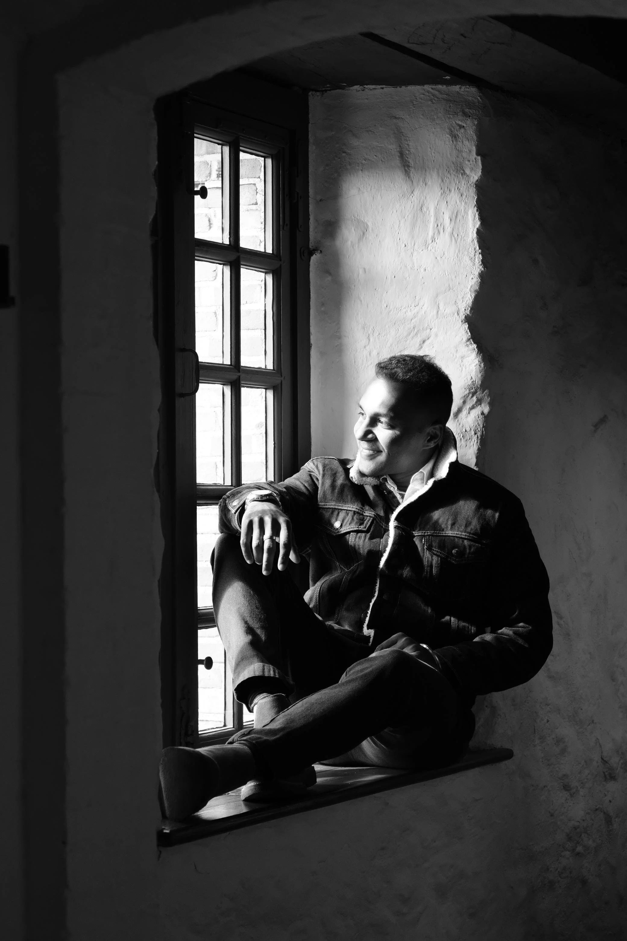 A young man sitting on a windowsill in a rustic, textured room, looking out with a smile.