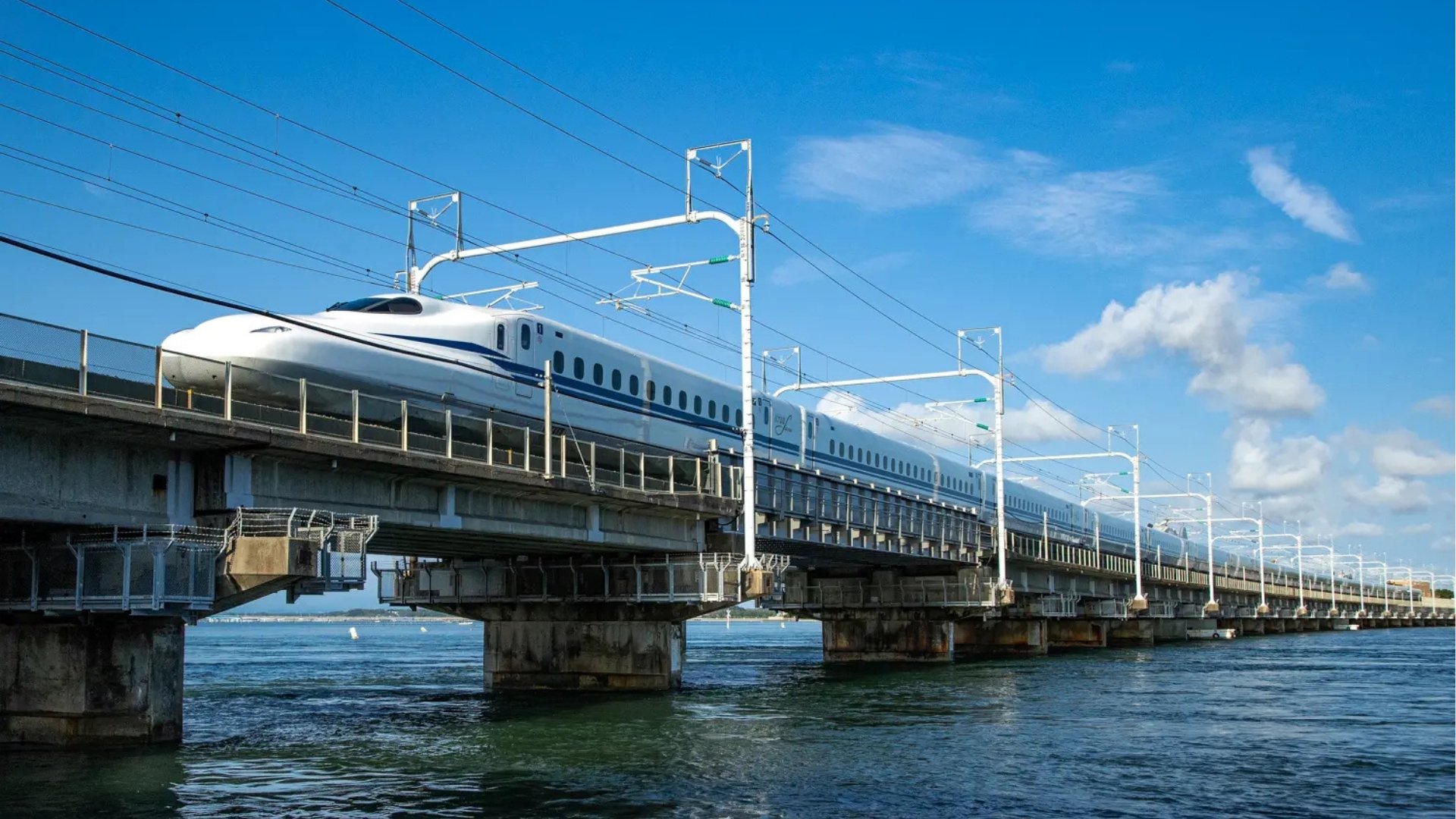 High-speed train traveling on a bridge over a body of water, with electrical wires overhead and a blue sky with scattered clouds in the background.