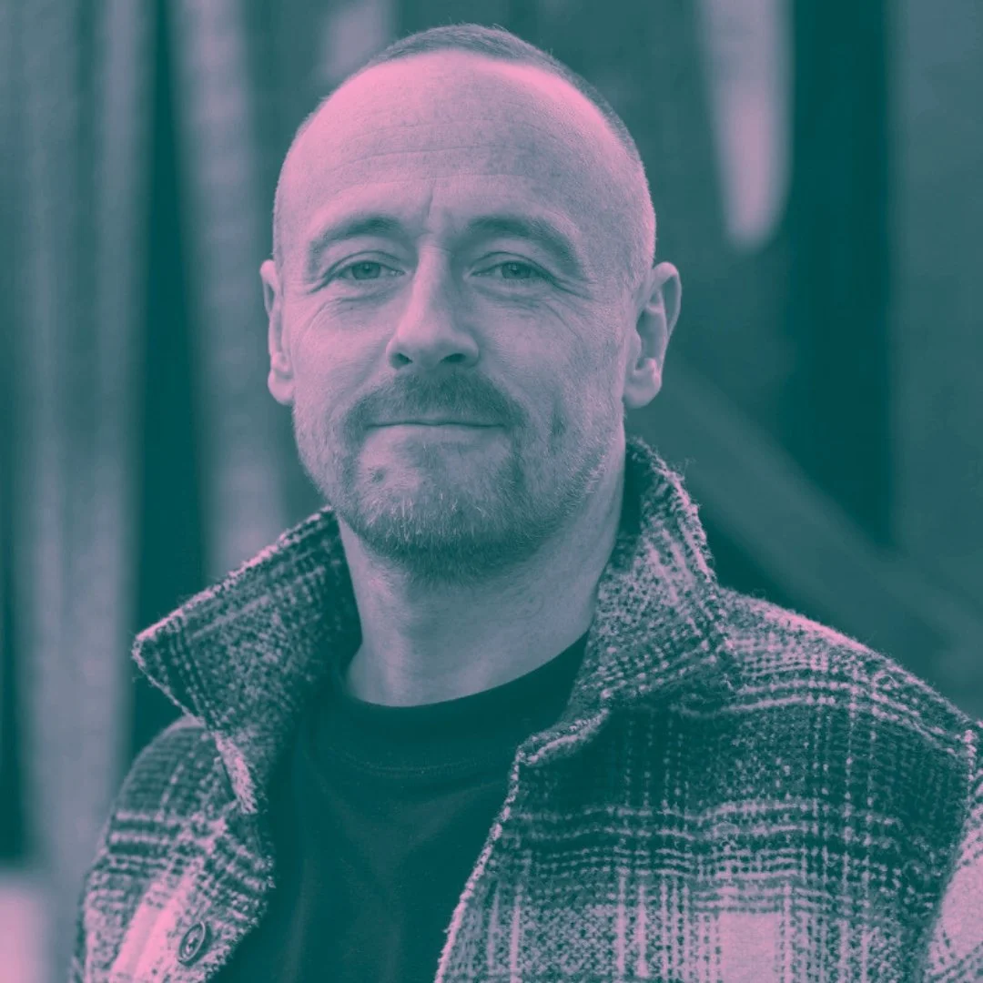 A man with a beard and short hair smiling outdoors, wearing a patterned jacket over a black shirt.