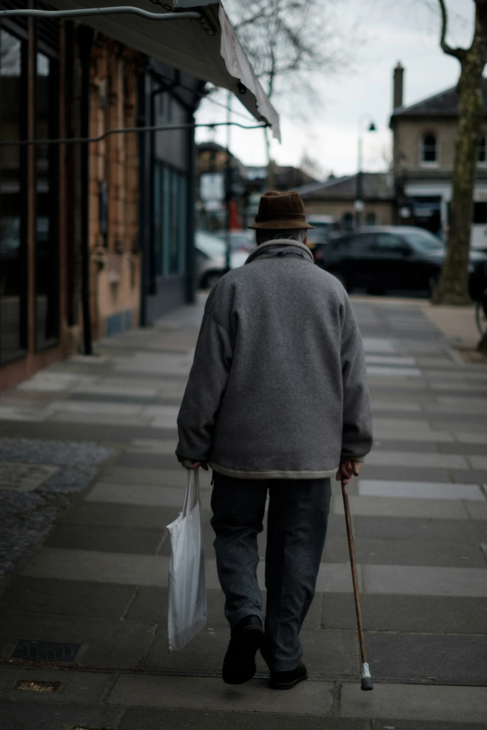 Un homme âgé marche avec une canne et porte un sac en plastique dans une main, vêtu d'un manteau gris et d'un chapeau, dans une rue urbaine pendant la journée.