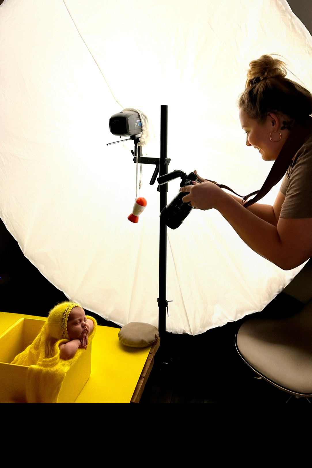 A professional photographer takes a photo of a baby with a yellow knitted hat, inside a yellow box, in a photography studio with a white backdrop.