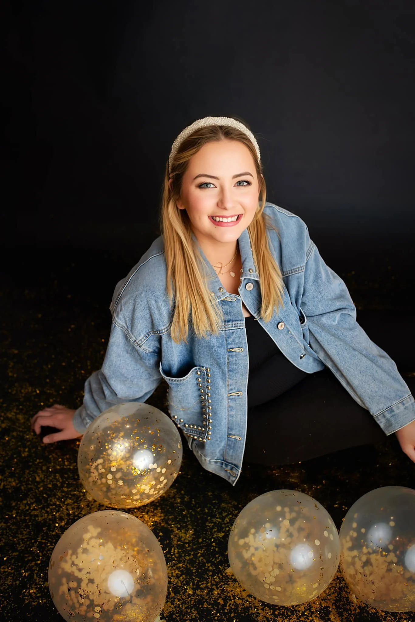 Young woman with blonde hair, wearing a denim jacket and a headband, sitting on a black surface with gold confetti and four transparent balloons with gold confetti inside, against a dark background.