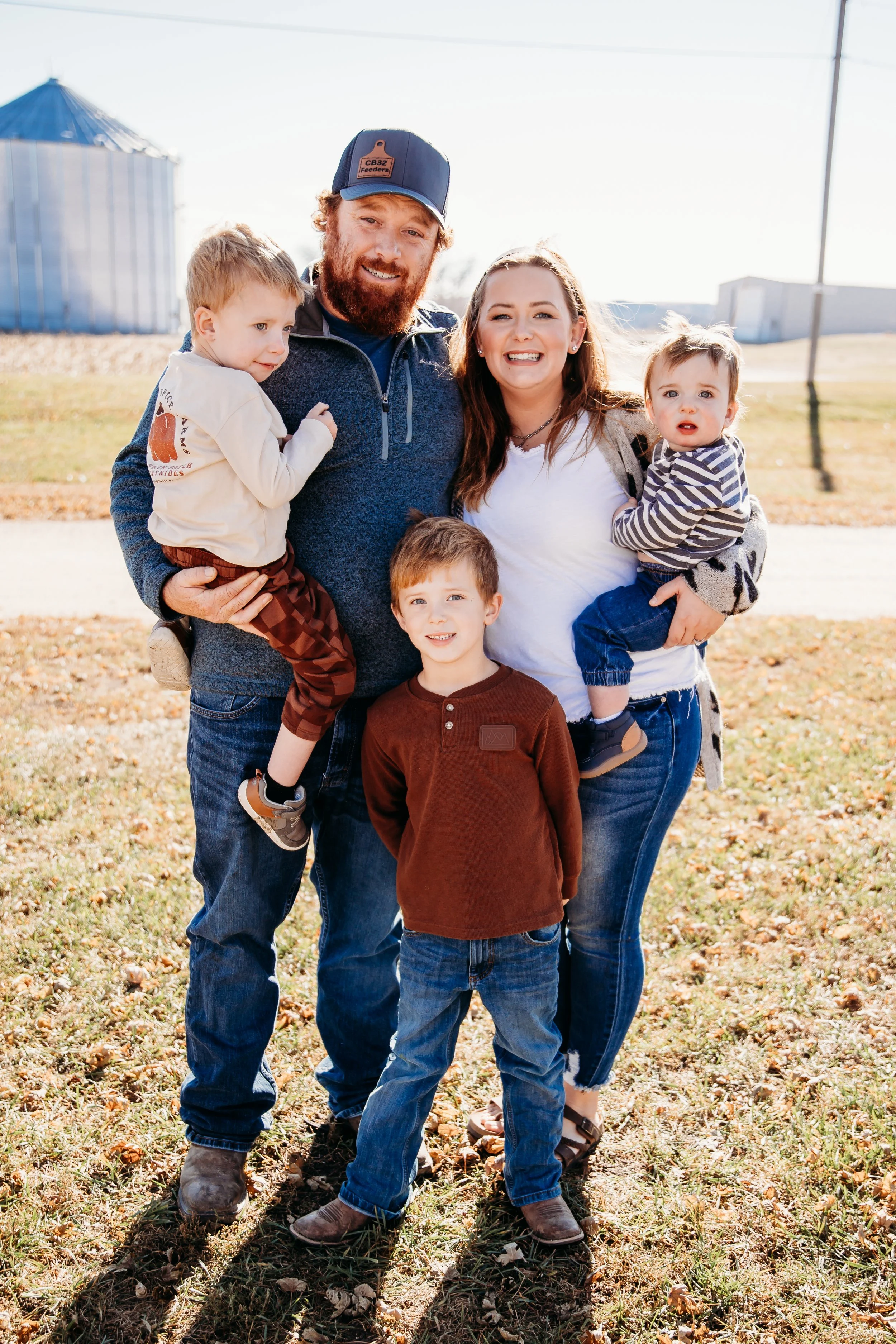 A family of five standing outdoors on a sunny day. The father has a beard and is wearing a baseball cap, the mother has long hair, and three children are with them, two being carried and one standing in front. They are all smiling and dressed casually.