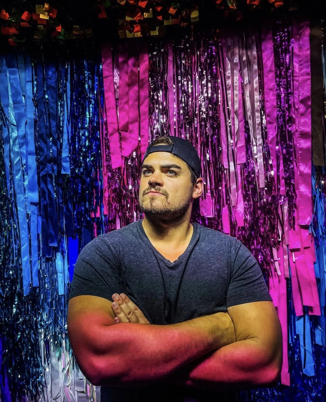 A man with a beard and dark hair wearing a backward baseball cap and a dark t-shirt, standing in front of a colorful background of pink, purple, blue, and black streamers with some shiny metallic decorations. He has his arms crossed and is looking to the side with a serious expression.