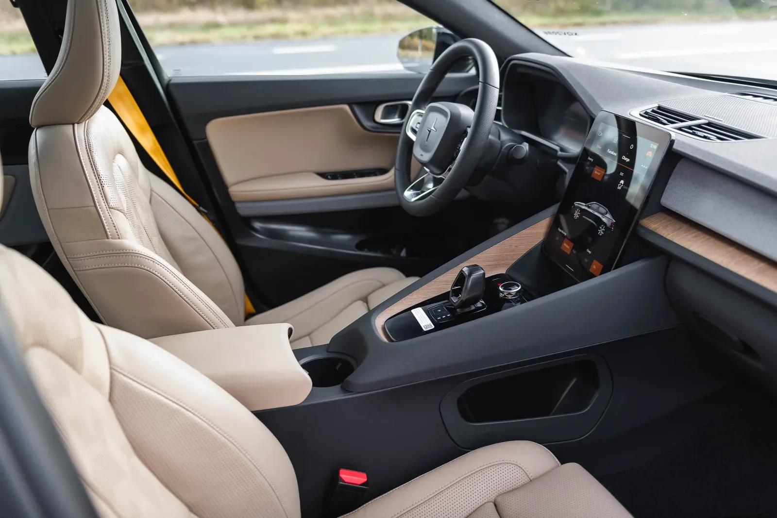 Interior of a modern car with beige seats, a steering wheel, and a dashboard with a large touchscreen display showing climate control options, parked on a highway.