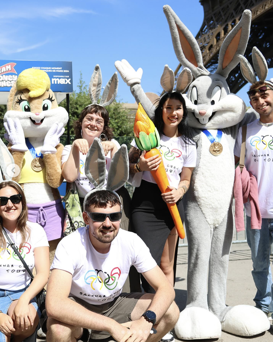 Group of people, including a person in a Bugs Bunny costume, posing outdoors with a bunny mascot, some wearing white T-shirts with Olympic logo, one holding an oversized carrot prop.