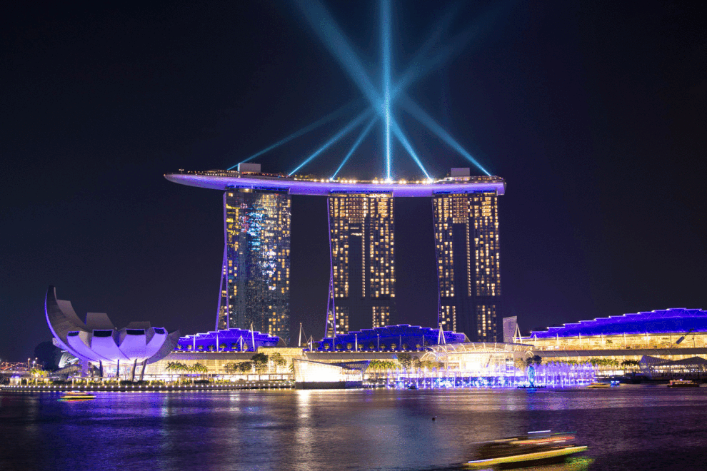 Nighttime view of Marina Bay Sands hotel in Singapore with laser light display projected from the rooftop, reflected on the water below.