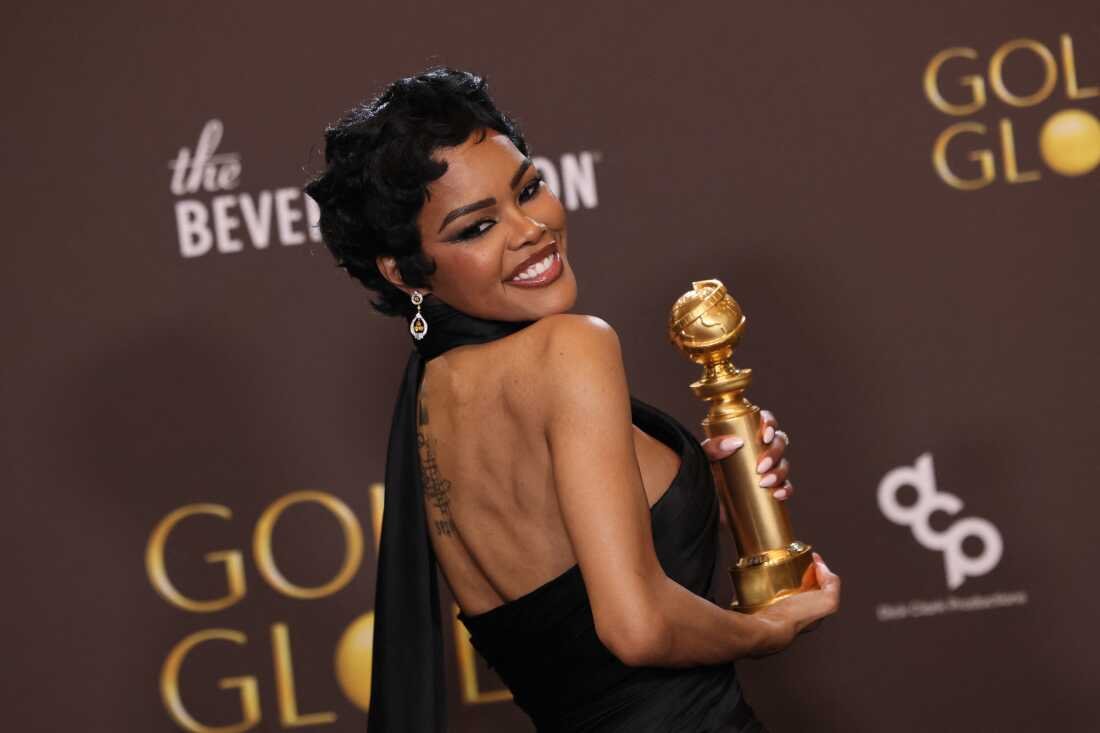 A woman with short curly hair and earrings, smiling and posing with a golden award statue at an event.