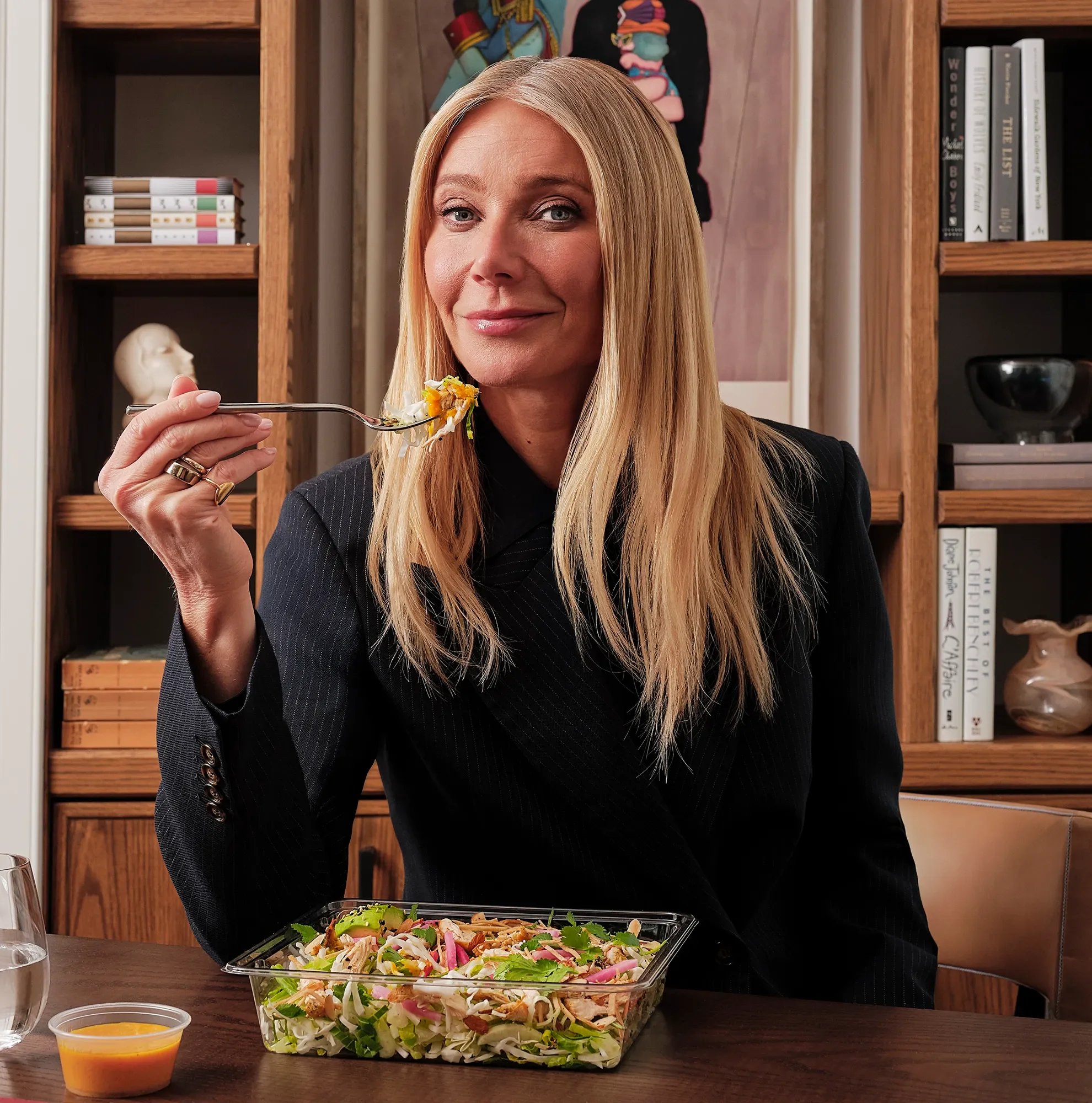 Woman with blonde hair and wearing a dark blazer, holding a fork with salad, standing behind a salad tray on a table, with bookshelves and artwork in the background.