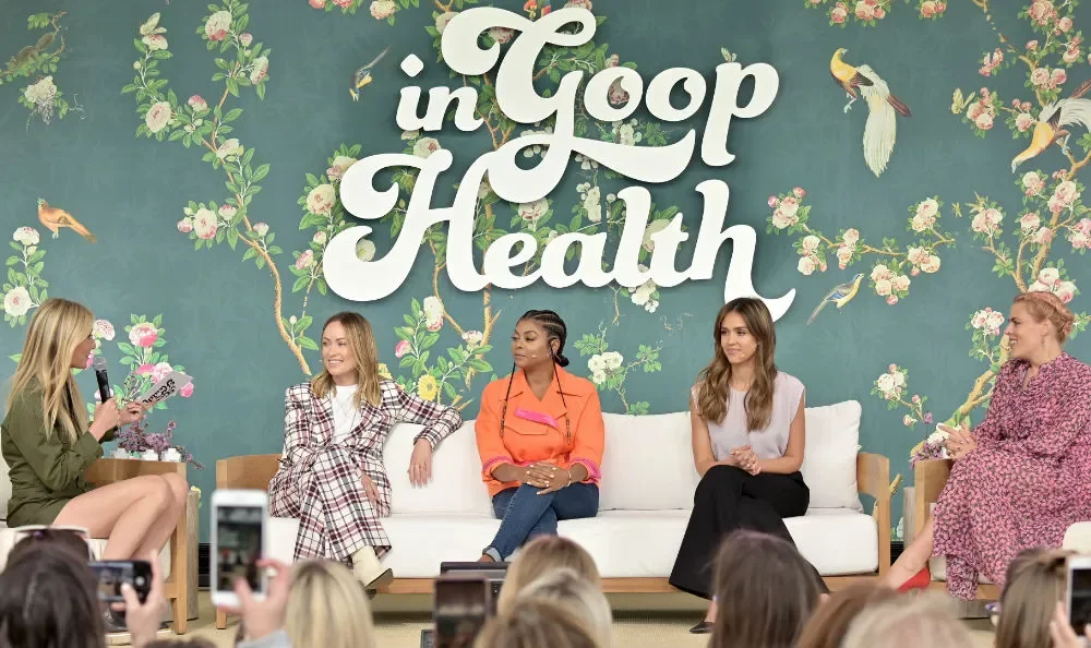 Five women sitting on a stage with a floral backdrop reading 'in Goop Health'; four women are engaged in conversation while one woman holds a microphone, and there is an audience taking photos in front.