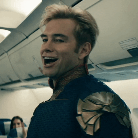 A smiling man with blond hair on an airplane, wearing a costume with a shoulder armor piece.