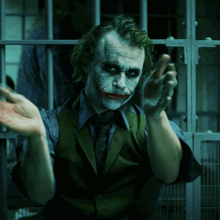 A man dressed as the Joker, wearing white face paint with black around the eyes and red lips, in front of metal bars, making a gesture with his hands.