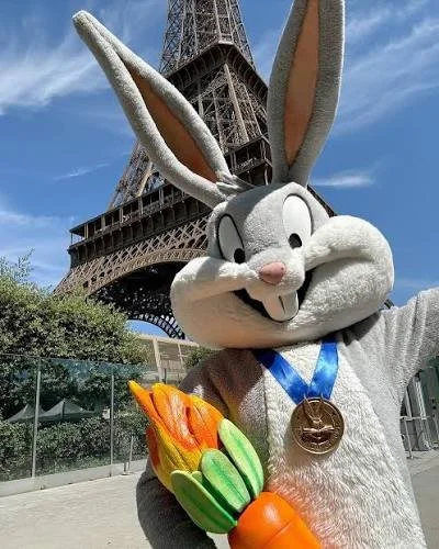 Bugs Bunny mascot with a gold medal and a carrot, standing in front of the Eiffel Tower.