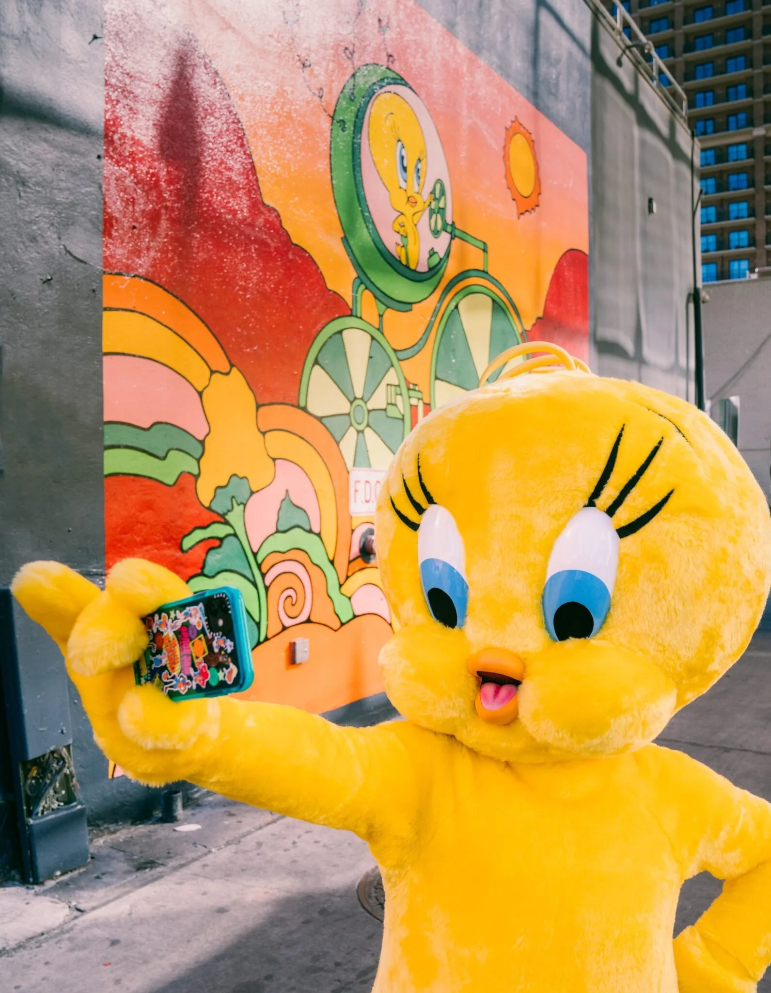 Close-up of a person in a yellow cartoon character costume taking a selfie with a smartphone, standing in front of a colorful mural and a building in the background.