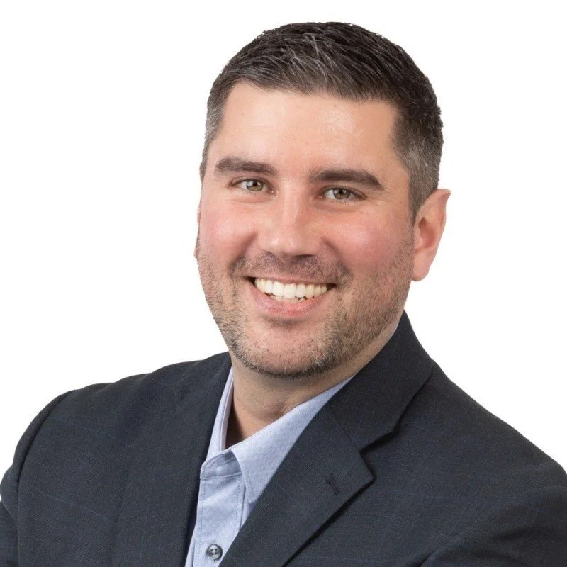 A young man with short dark hair, light skin, and a gentle smile, wearing a dark suit jacket and a light blue shirt, posing against a white background.