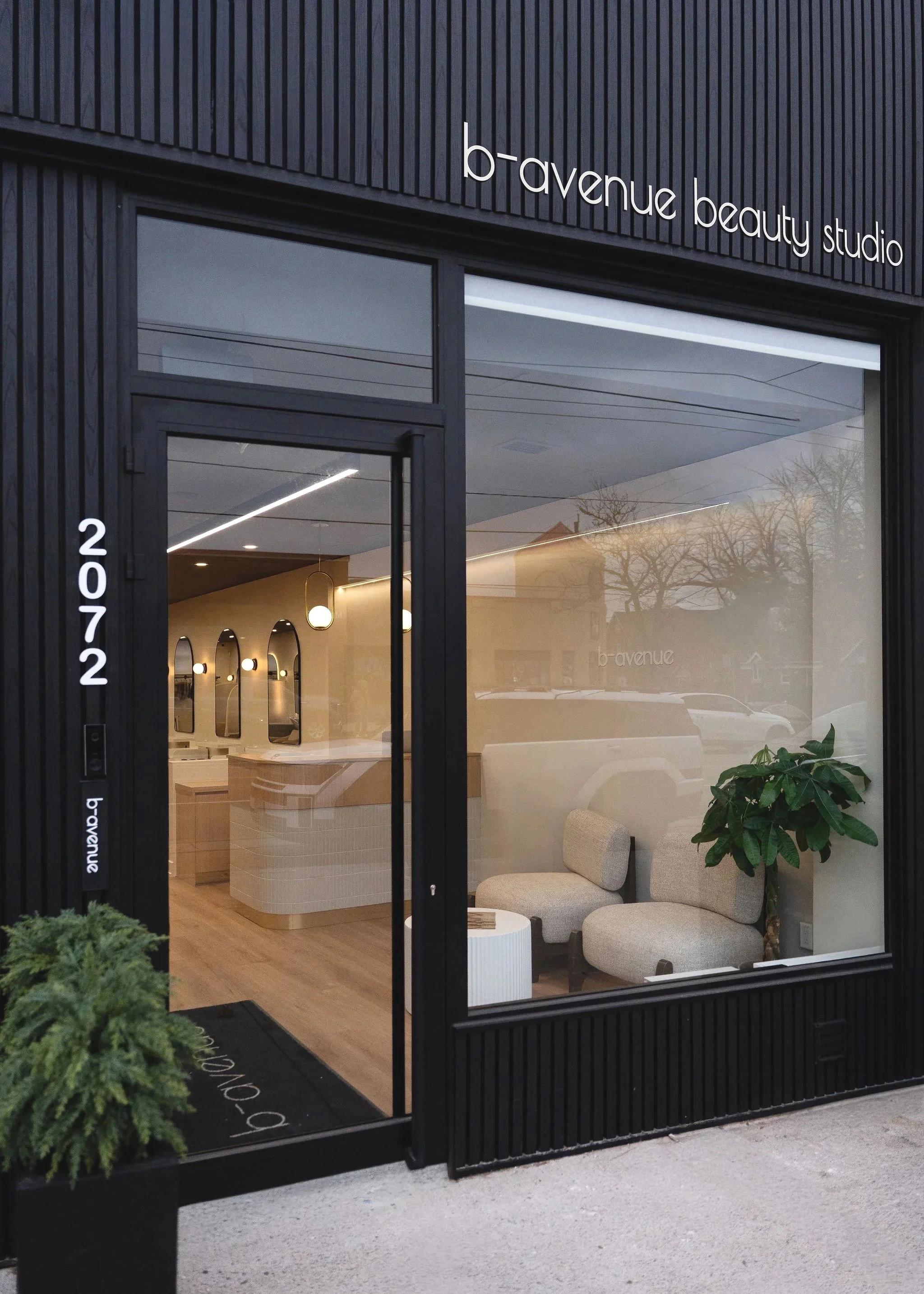 Exterior view of b-avenue beauty studio with black facade and large glass window showing a modern, minimalist interior with seating area, potted plant, and reception desk.