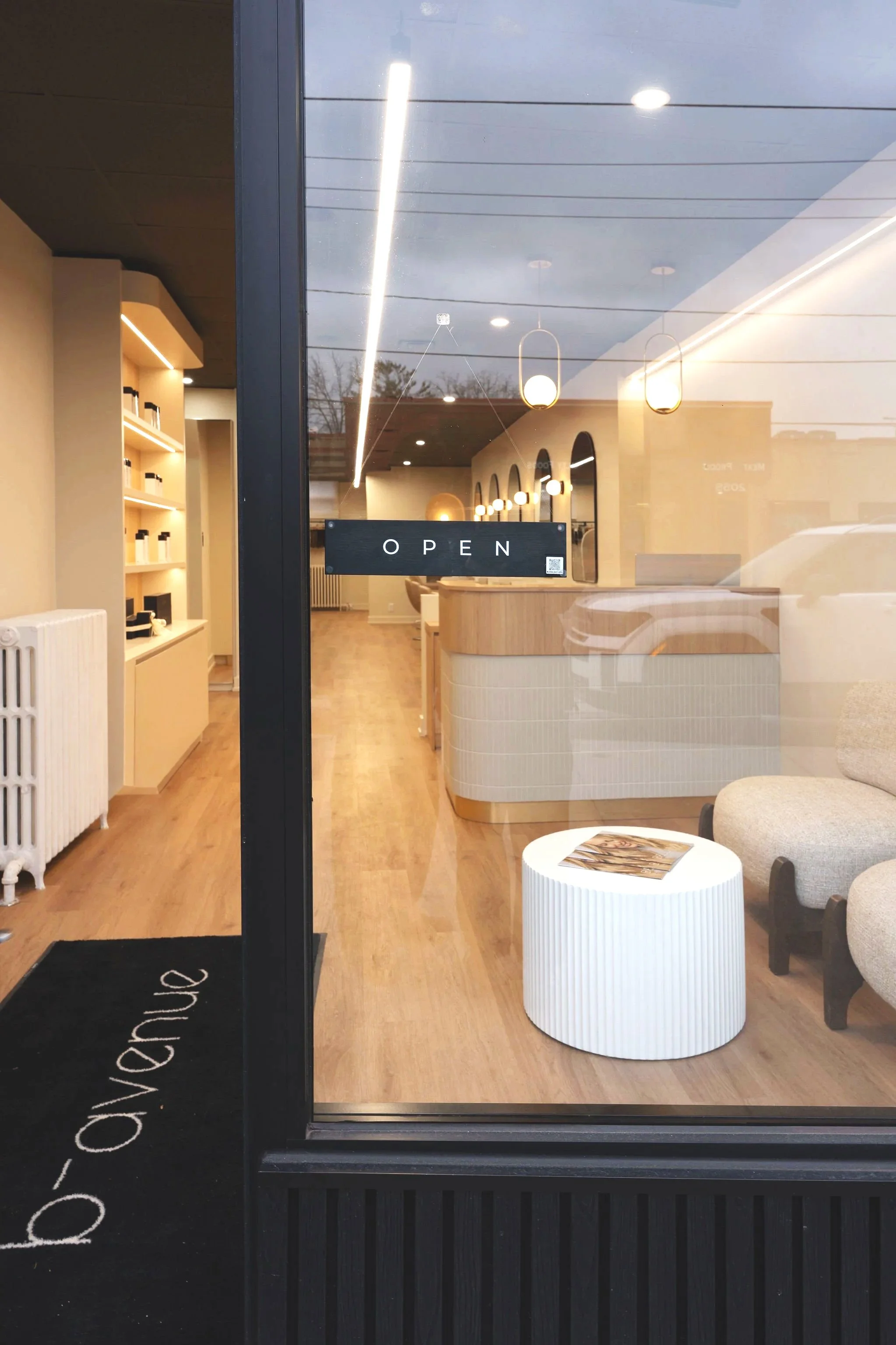 View through glass door of a modern, minimalist interior space with a black 'OPEN' sign on the door, white walls, warm wooden flooring, and contemporary furniture, including a white cylindrical side table and beige chairs.