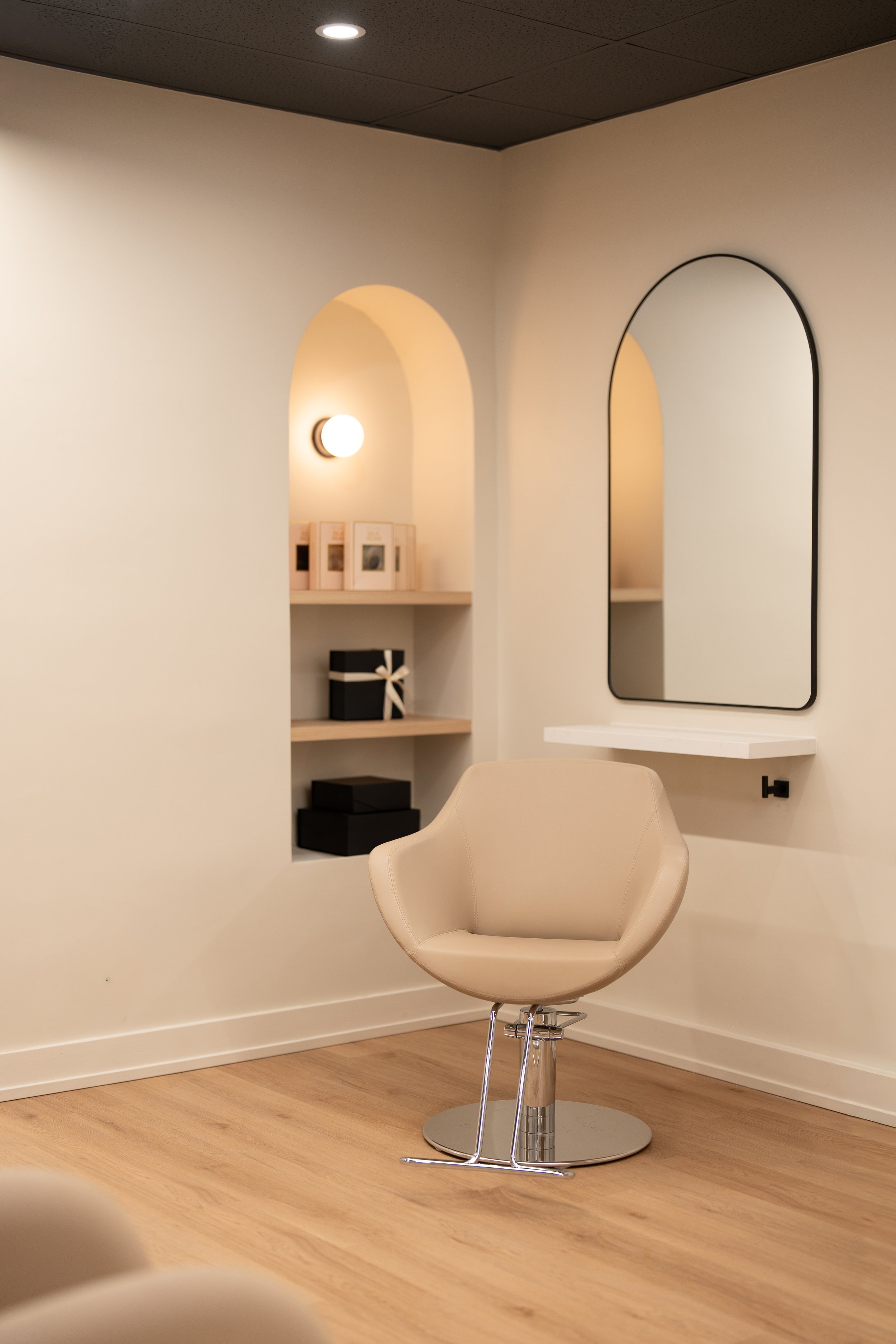 A beige salon chair in front of an arched mirror on a wall, with built-in shelves holding black boxes and framed photos, inside a room with wooden flooring and a black ceiling.
