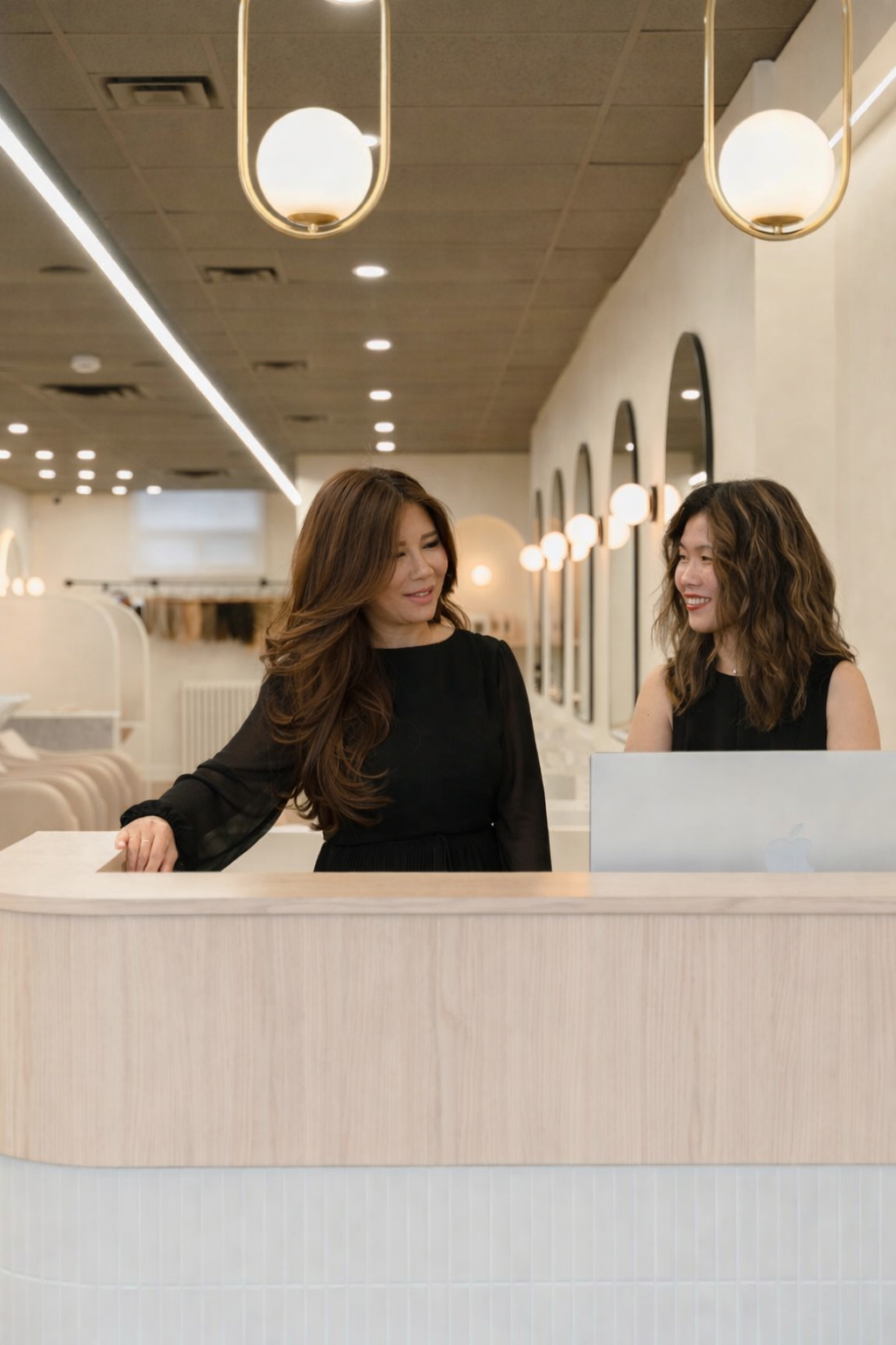Two women smiling and talking at a reception counter with a laptop in a modern, well-lit space.