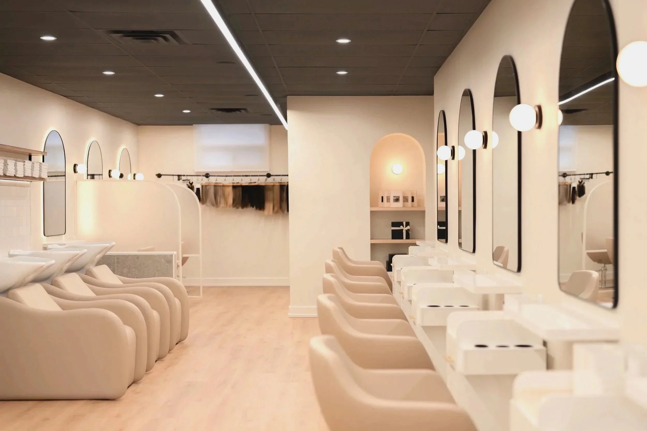 Empty salon with hair washing stations on the left and stylist stations with mirrors and chairs on the right, in a modern, well-lit interior.
