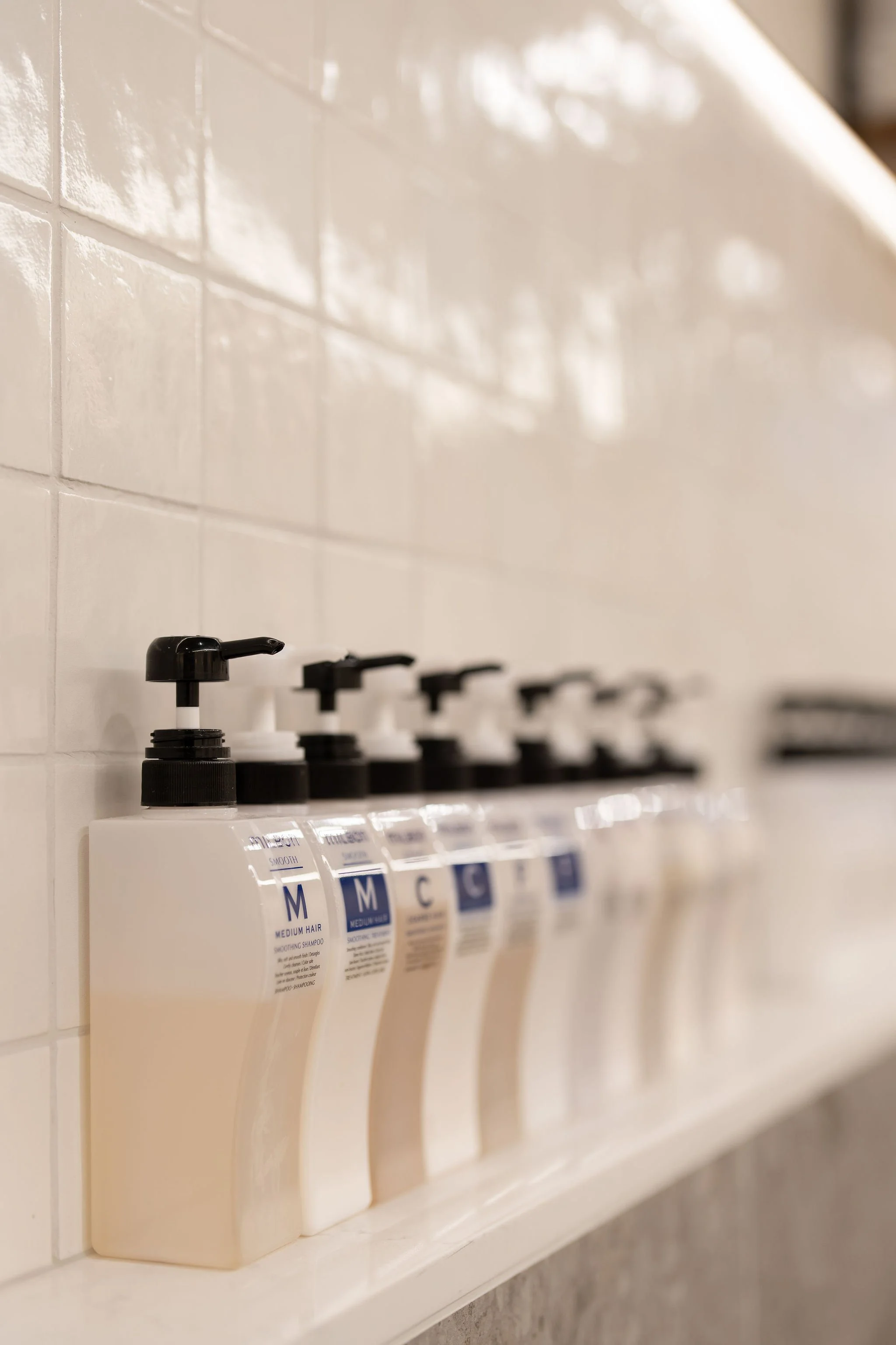 Six white bottles with black pump dispensers lined up on a white shelf against a tiled wall, likely shampoo and conditioner bottles in a bathroom or salon.
