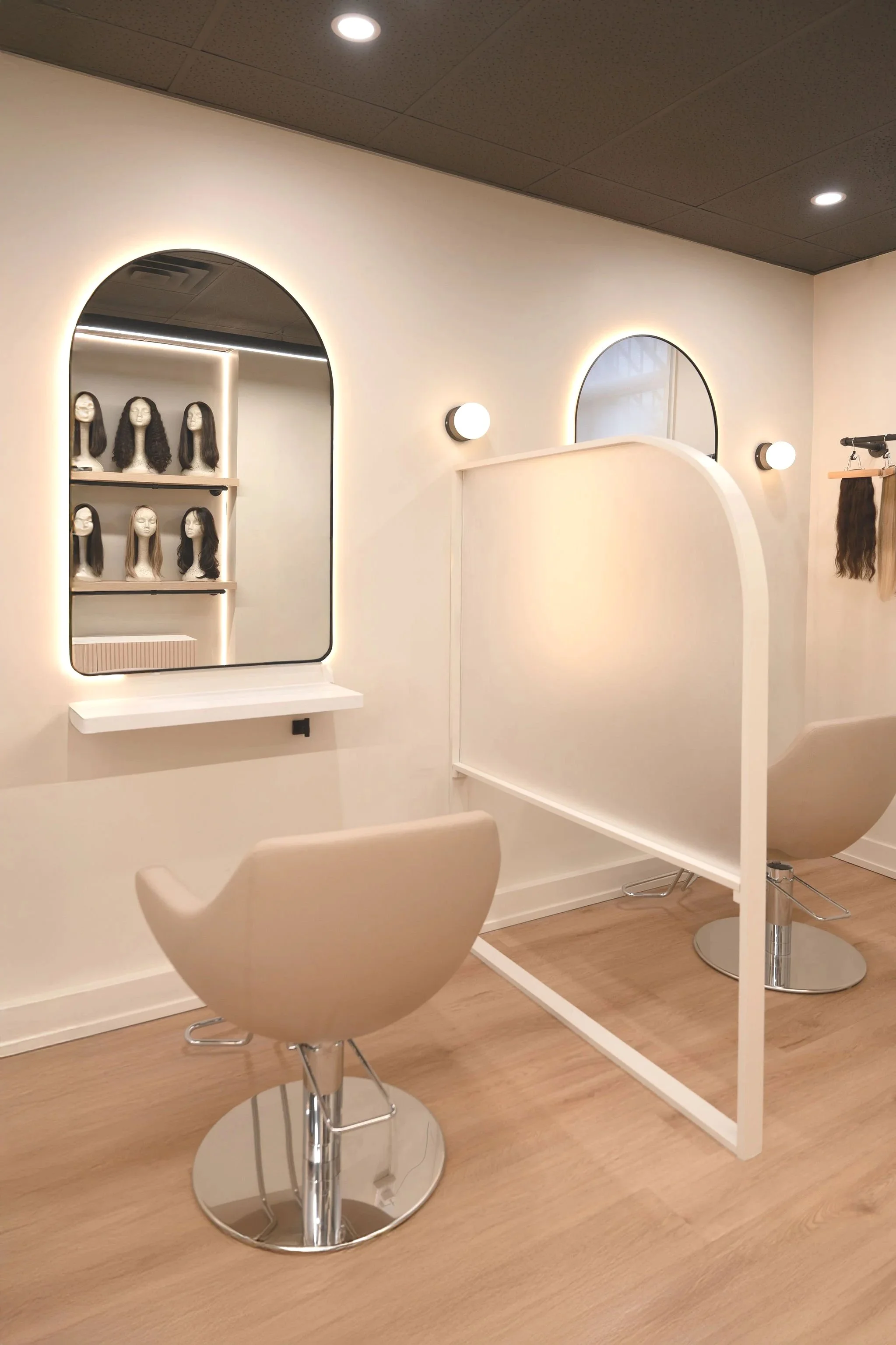 A modern hair salon with two styling chairs, a large illuminated mirror, and a display of mannequin heads with various hairstyles on shelves.