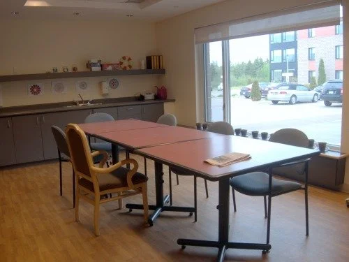 Meeting room with a large table surrounded by chairs, a window with a view outside, and a kitchenette with shelves and cabinets in the background.