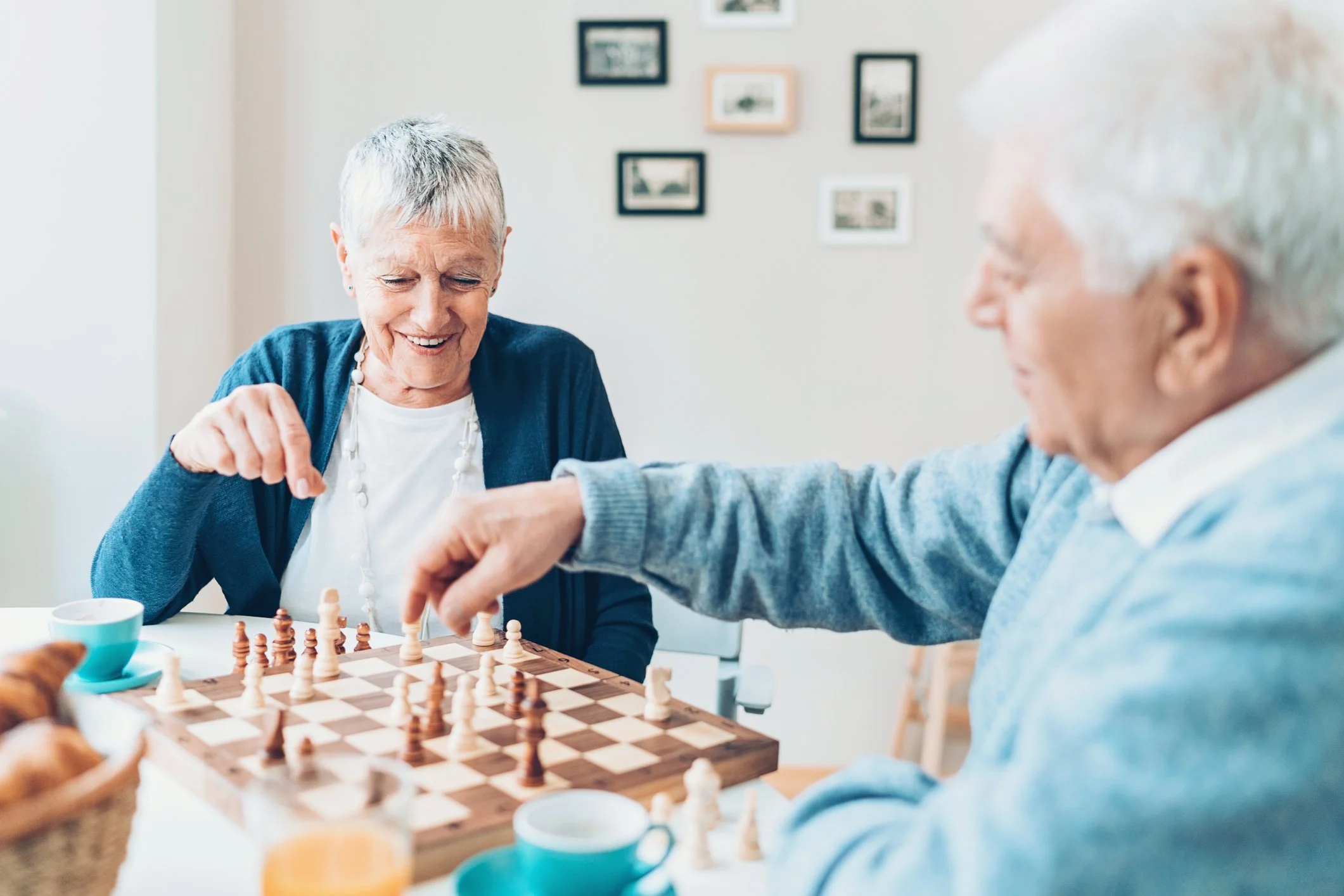 An elderly woman and man are playing chess at a table indoors, smiling and enjoying their game with cups and a basket of croissants nearby.