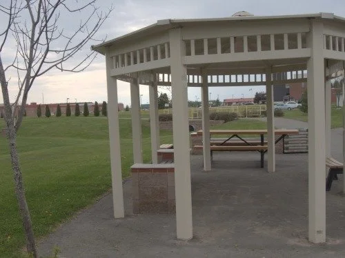 A white wooden gazebo with a picnic table inside, located in a park with grass and trees.