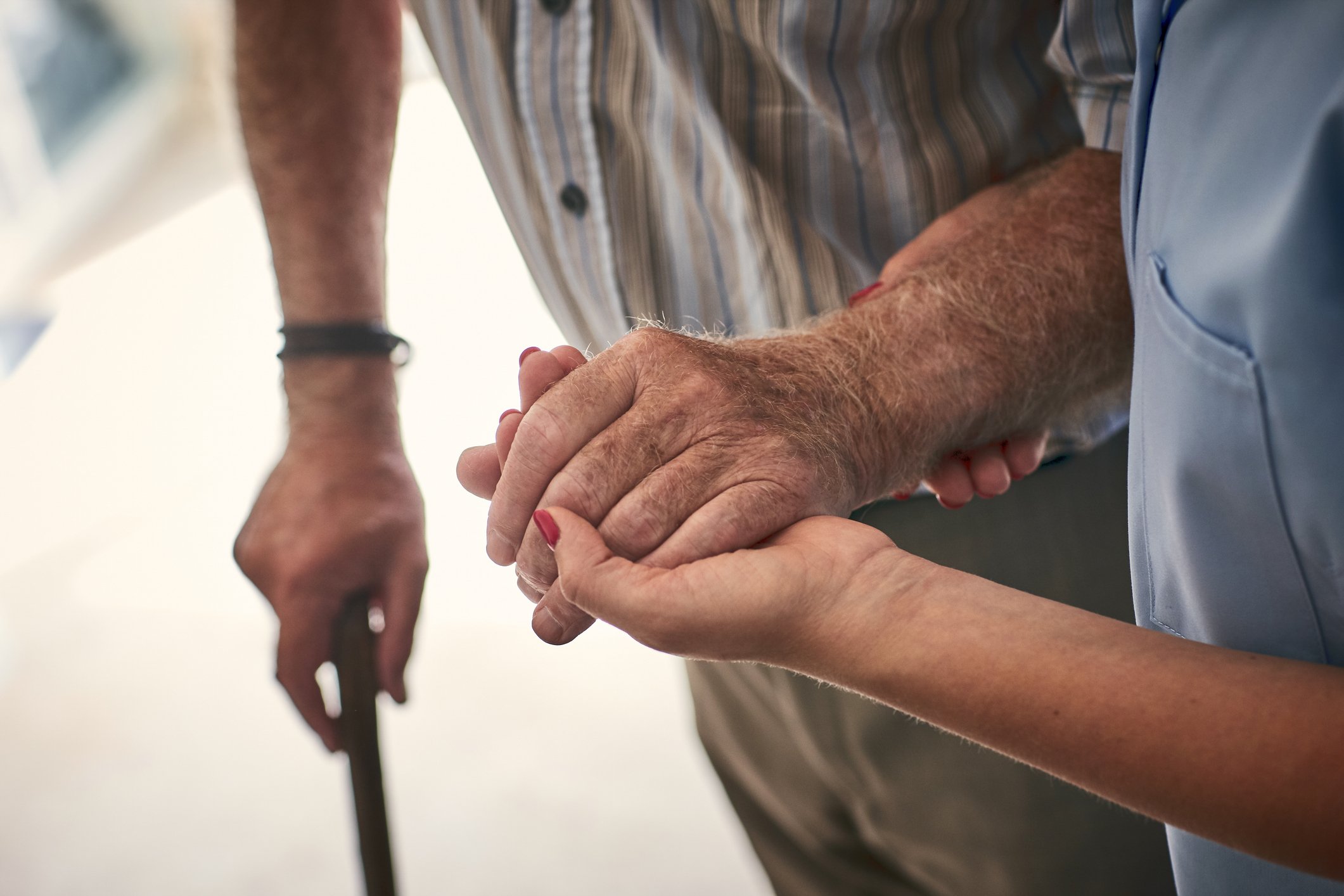 Close-up of a person helping an elderly person walk by holding their hand, with another person in the background.